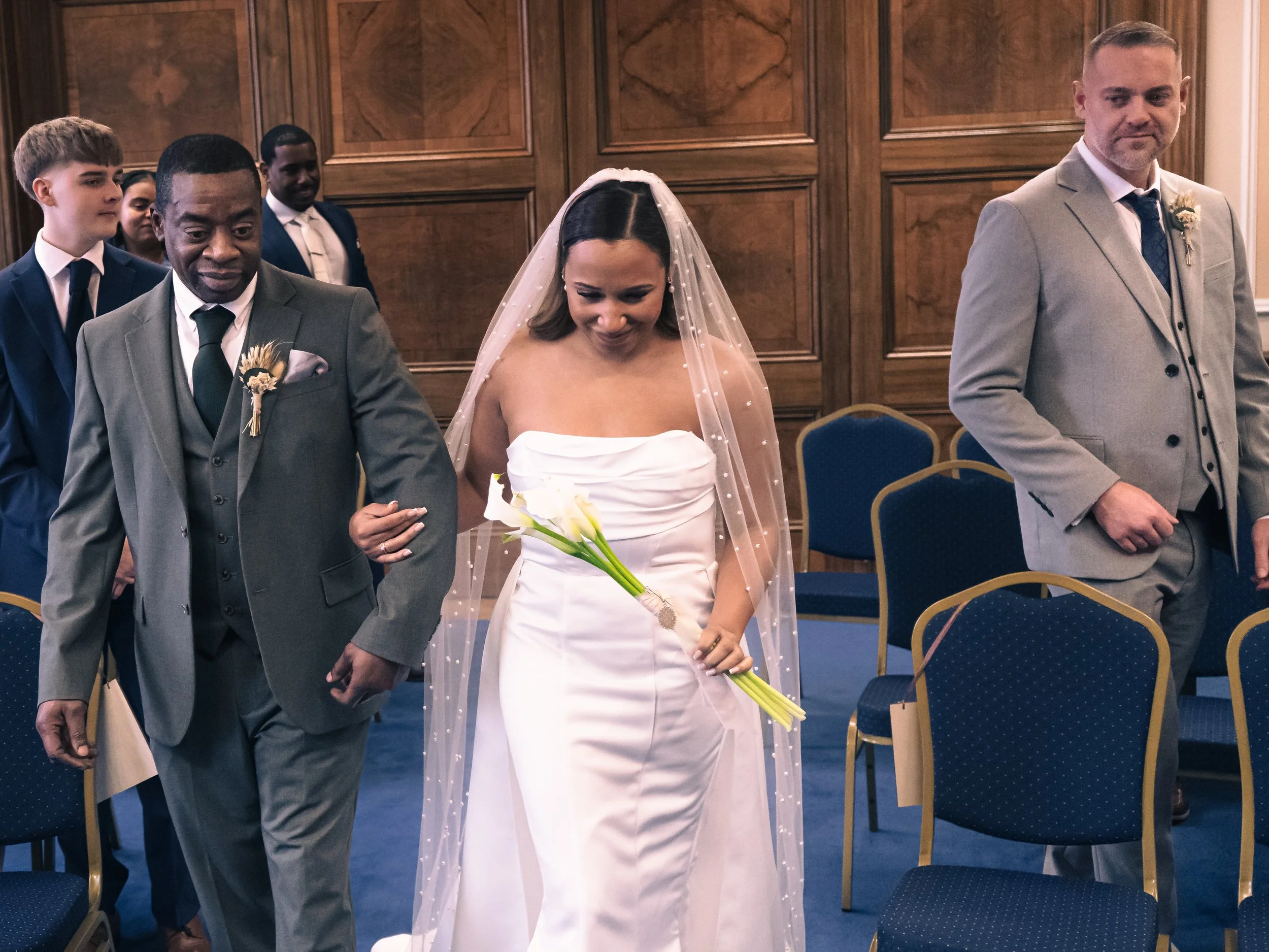 A bride in a white wedding dress and veil holding a bouquet of calla lilies, walking down an aisle with two men, one on each side, in a wood-paneled room with blue chairs.