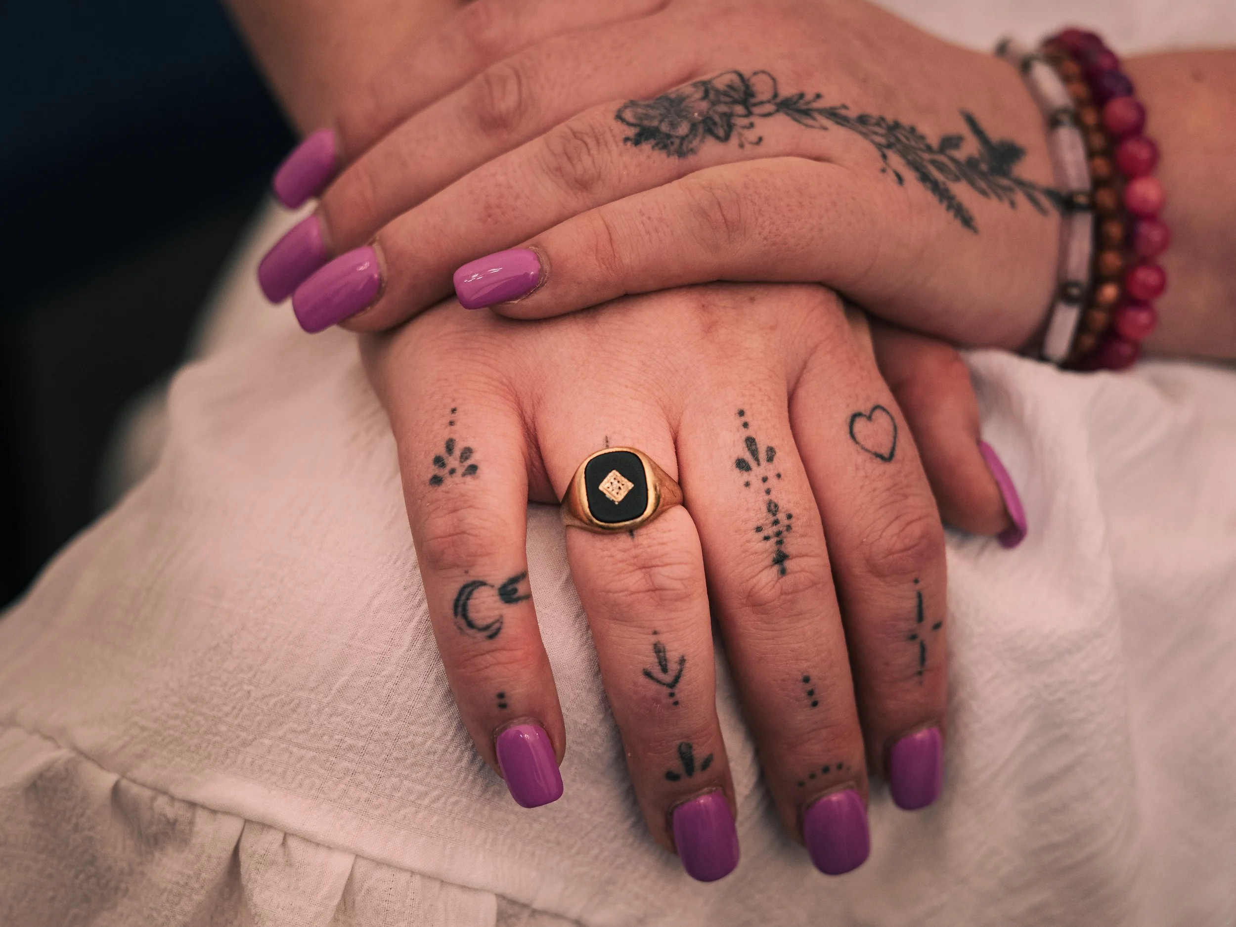 Close-up of two hands with purple painted nails, overlapping. The hands have multiple small tattoo designs, including hearts, moons, arrows, and floral motifs. One hand has rings, and the other has beaded and bracelet accessories.