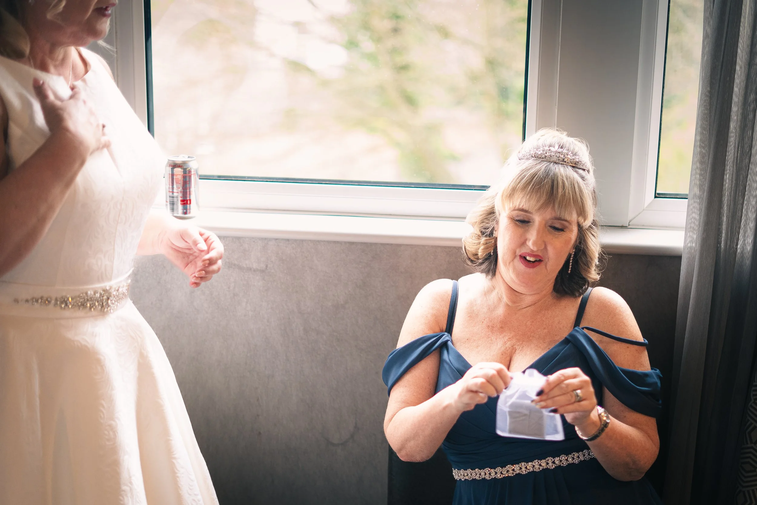 A woman in a blue dress sitting by a window, opening a small gift or card, with another woman in a white dress standing nearby and a can of soda on the windowsill.