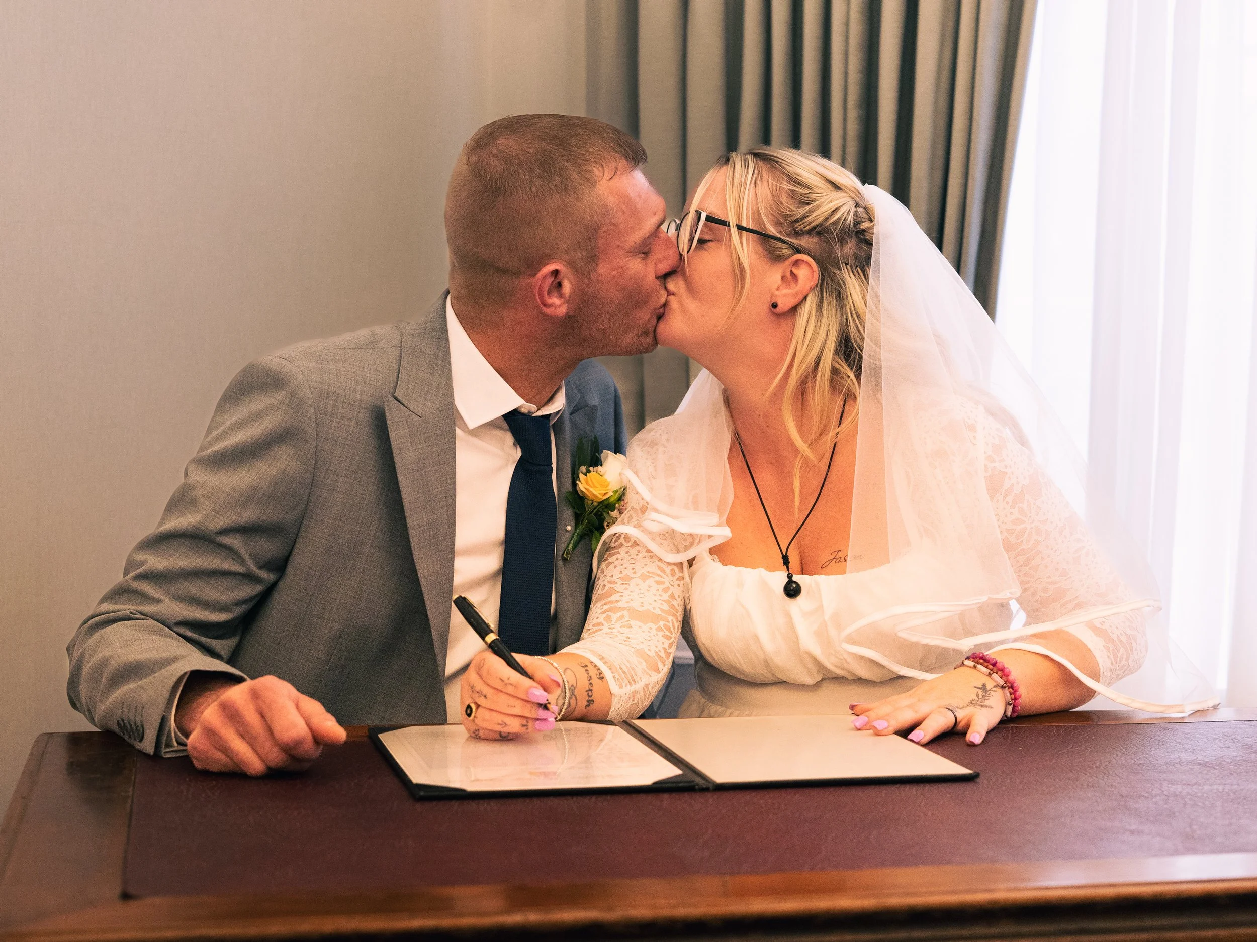 A couple in wedding attire kissing as they sign a marriage certificate at a table inside a room with curtains and daylight shining through the window.