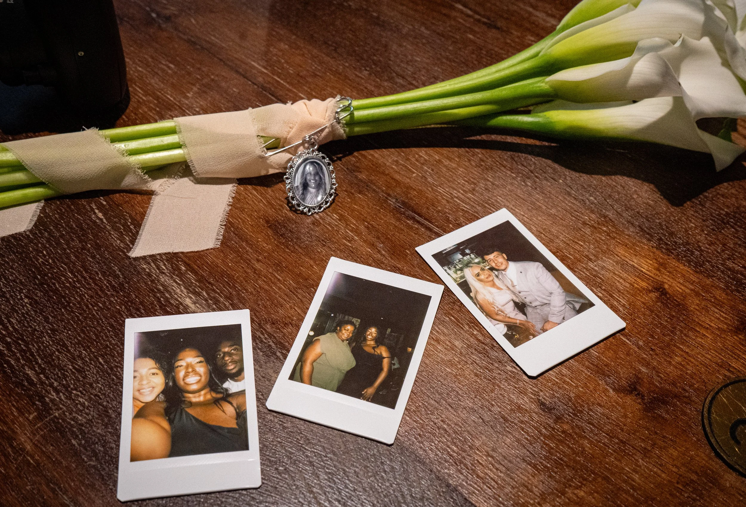 A wooden surface with three instant photographs, a bouquet of white calla lilies wrapped in beige ribbon, and a silver necklace with a pendant depicting a woman's face.