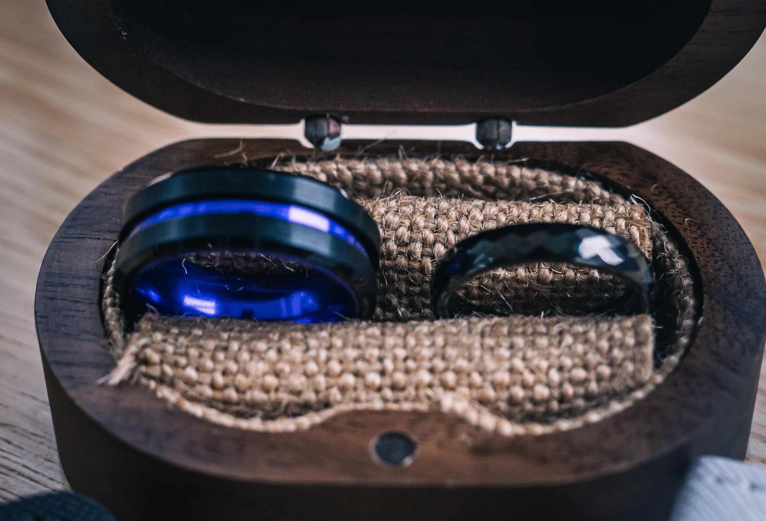 Open wooden case containing a pair of black over-ear wireless headphones with blue accents, resting on a woven fabric interior.