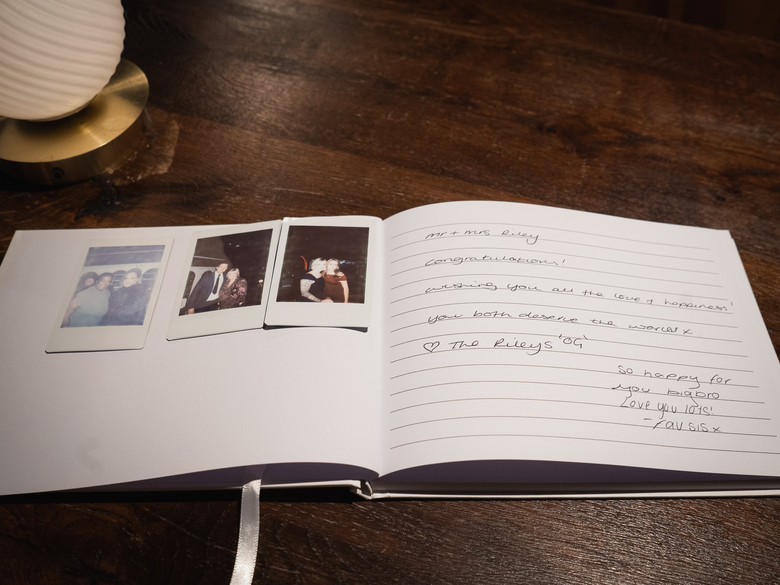 Open guestbook with three polaroid photos on left page and handwritten message on right page, on a wooden table with a lamp in the background.