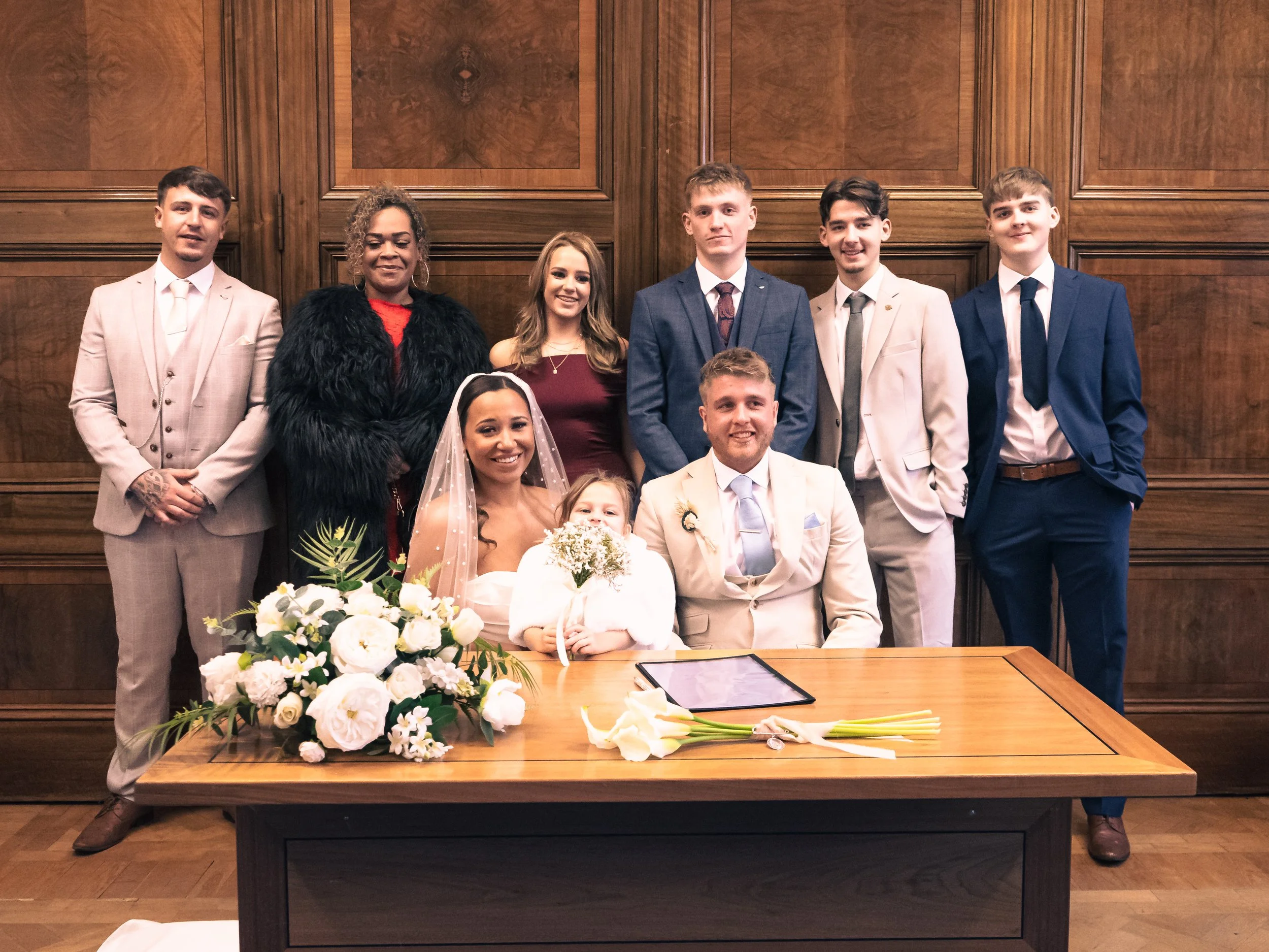 A wedding group photo featuring a bride and groom seated in front of a wooden wall, surrounded by eight friends or family members. The bride holds a bouquet and wears a veil, while the groom wears a light-colored suit. A floral arrangement and a docu