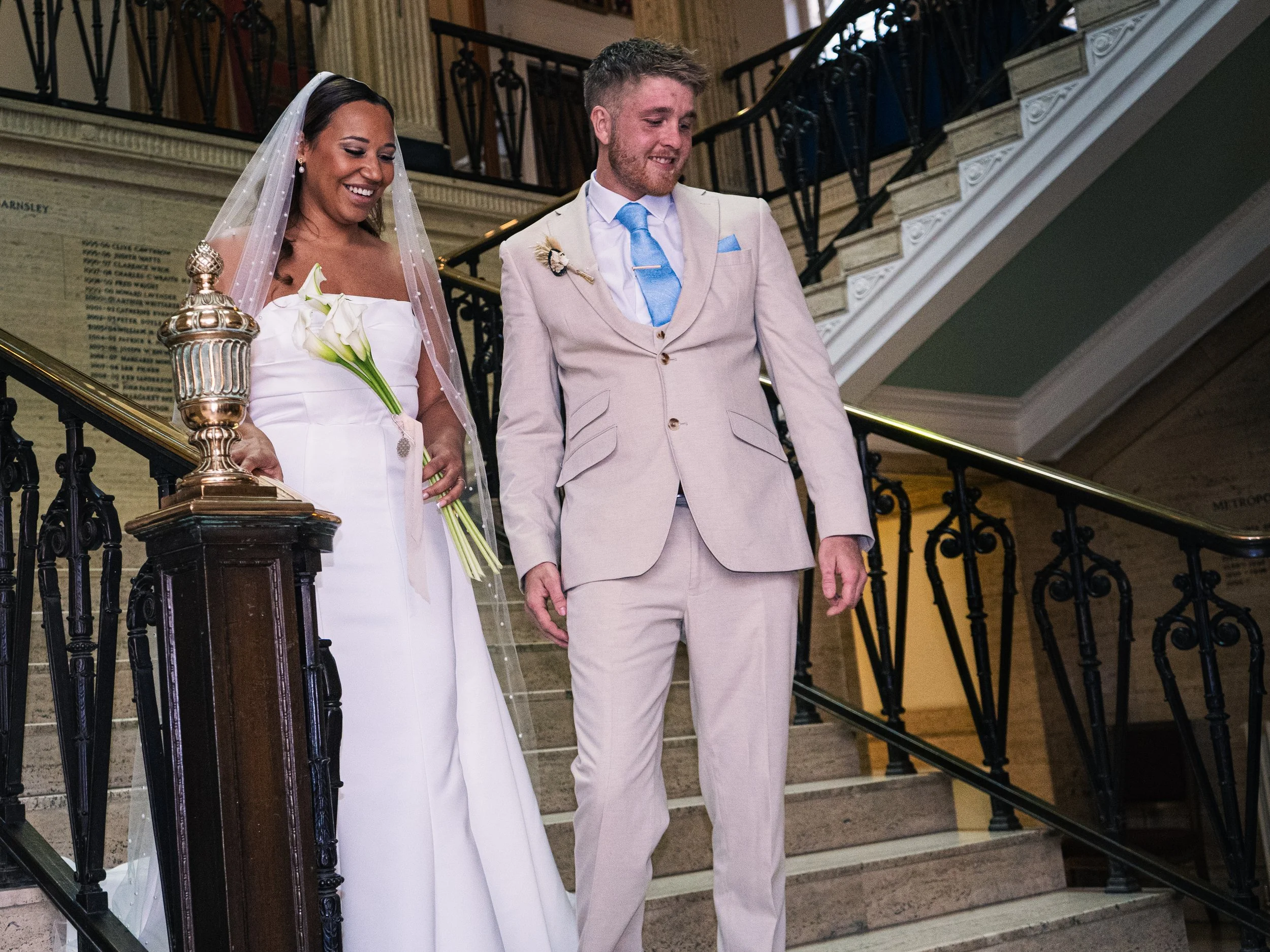 A bride in a white wedding dress with a veil holding a white calla lily bouquet, standing beside a groom in a light beige suit with a blue tie, on a staircase inside a building with ornate railings.