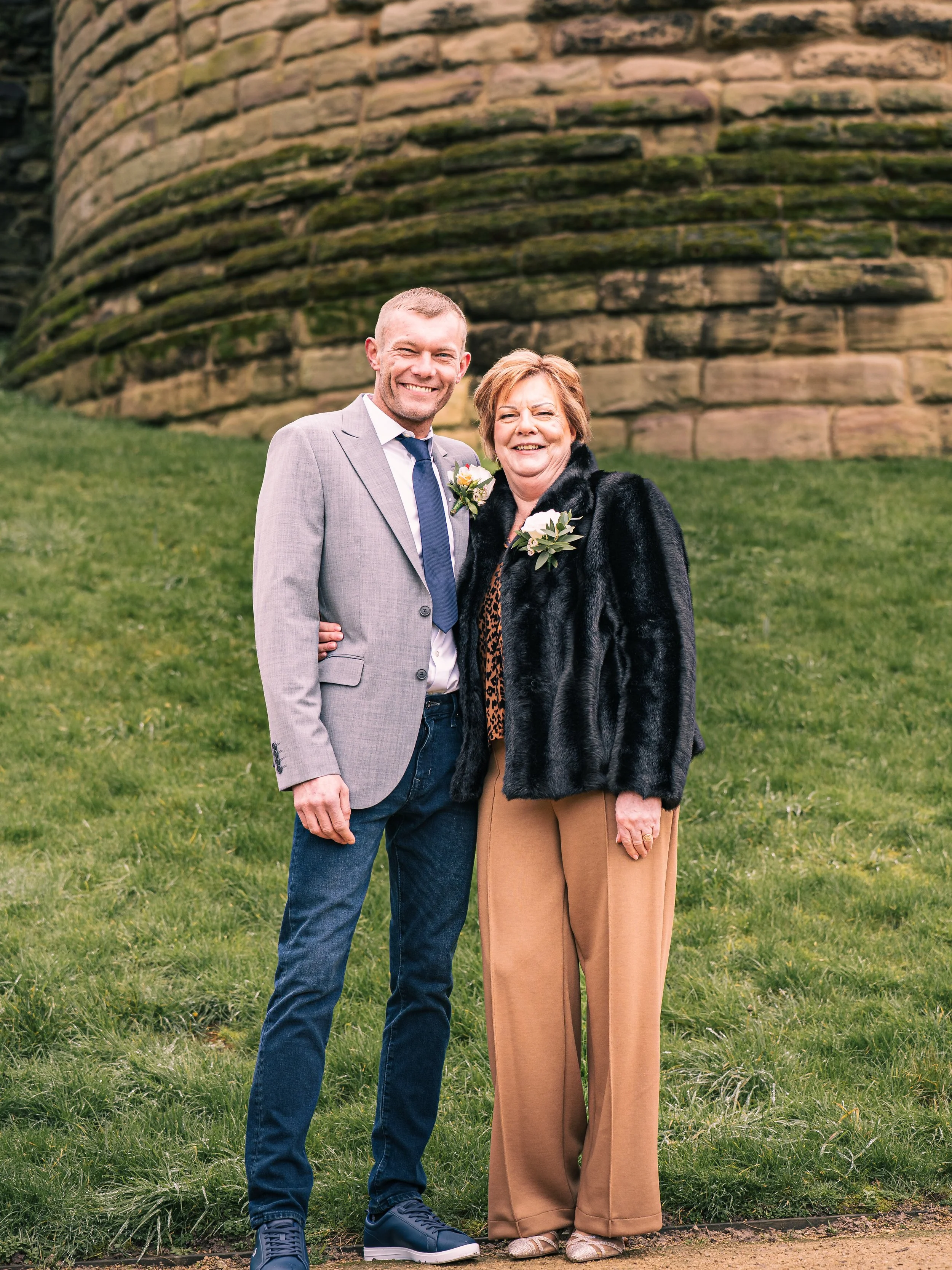 A smiling man and woman standing side by side on grass in front of a stone structure, dressed in formal attire with boutonnieres and corsages, indicating a special event like a wedding.