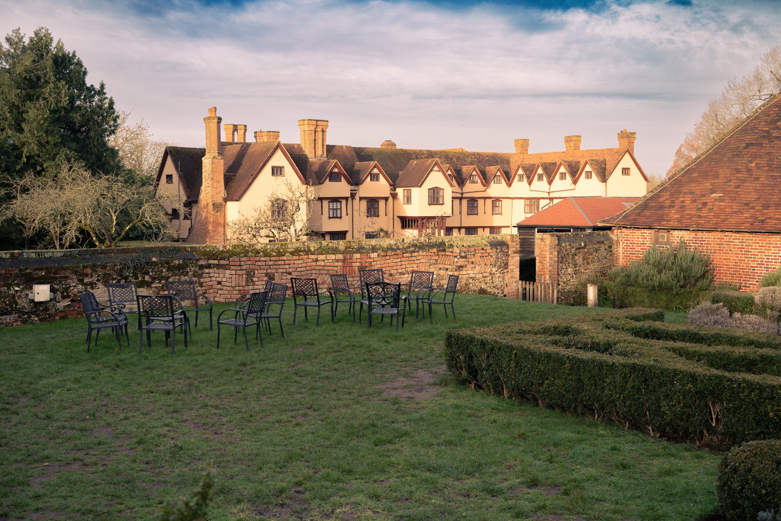 An outdoor garden area with ten black metal chairs arranged in a semi-circle on a grassy lawn, surrounded by a brick wall and historic-looking buildings with wood-framed windows and chimneys in the background, under a partly cloudy sky.