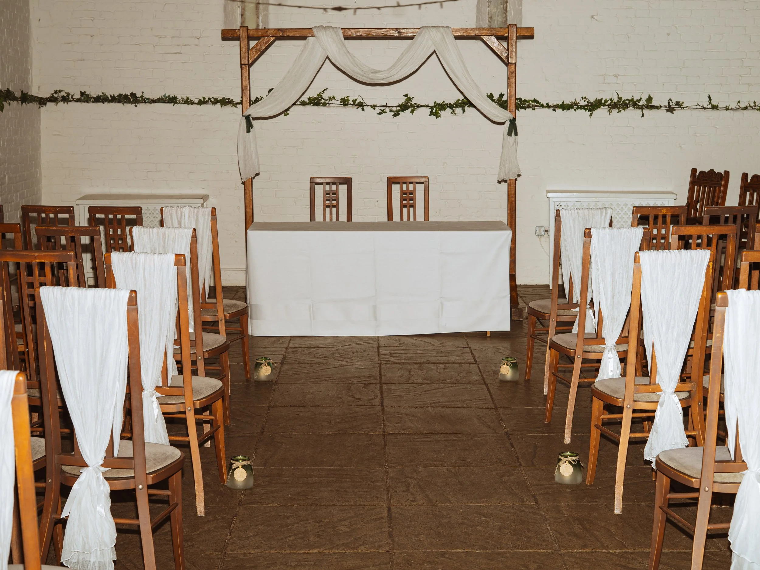 An indoor wedding ceremony setup with a table and two chairs at the front, surrounded by chairs with white fabric drapes on the backs, and small lanterns along the aisle. A fabric-draped wooden frame is above the table, against a white brick wall dec