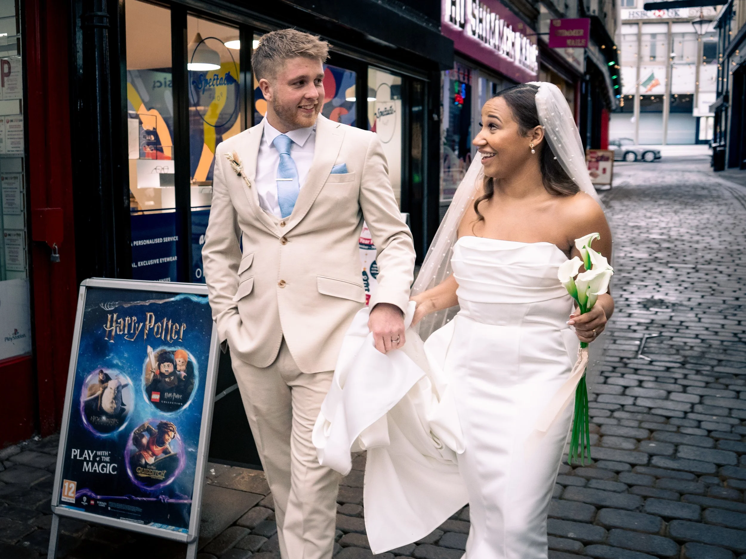 A newlywed couple walking on a cobblestone street; the groom is in a cream suit and the bride is in a white strapless wedding dress holding calla lilies; they are smiling at each other.