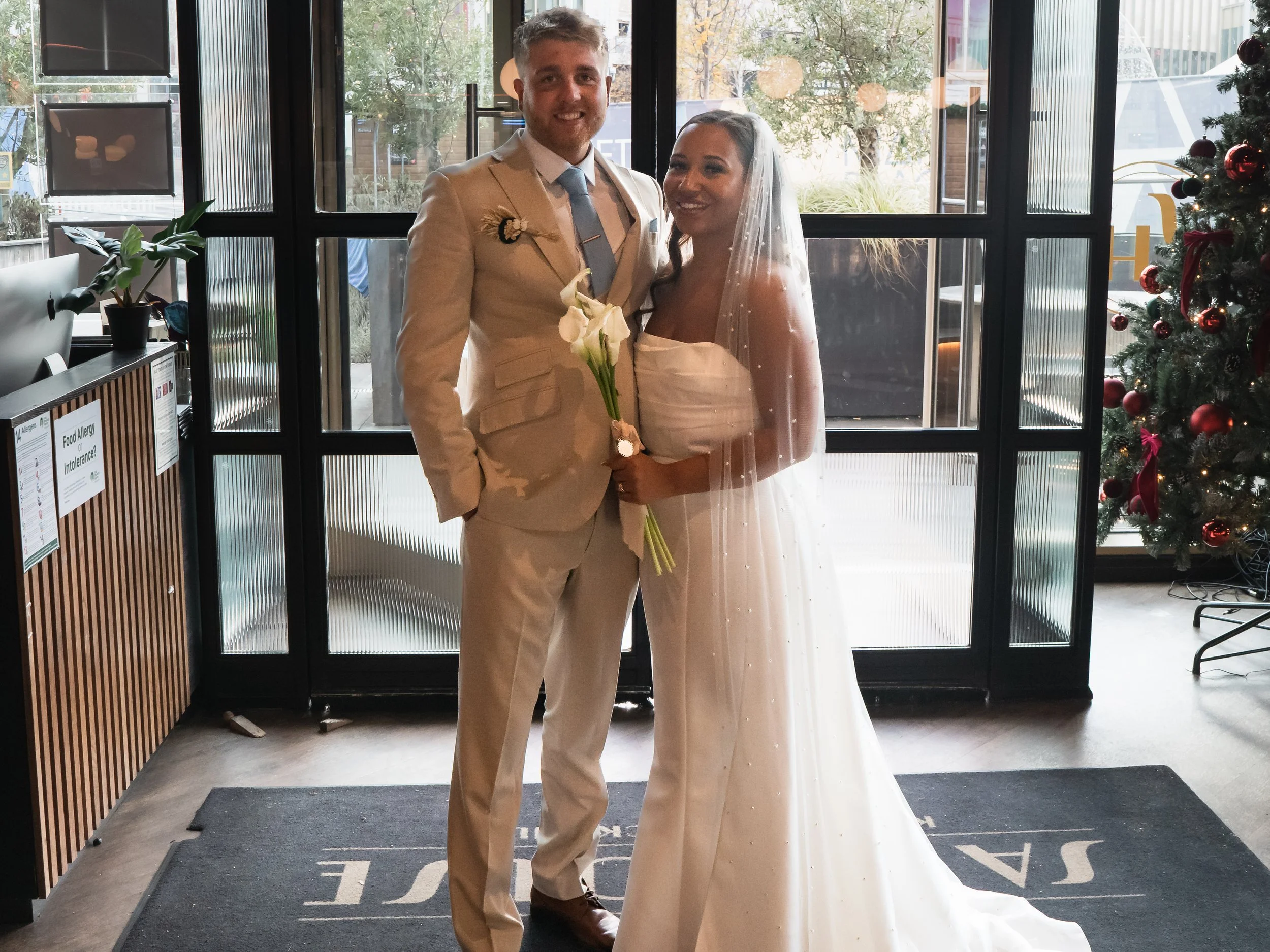 A couple dressed in wedding attire standing inside a building, with a Christmas tree decorated with red ornaments and ribbons on the right side and large windows behind them, holding a bouquet of white calla lilies.