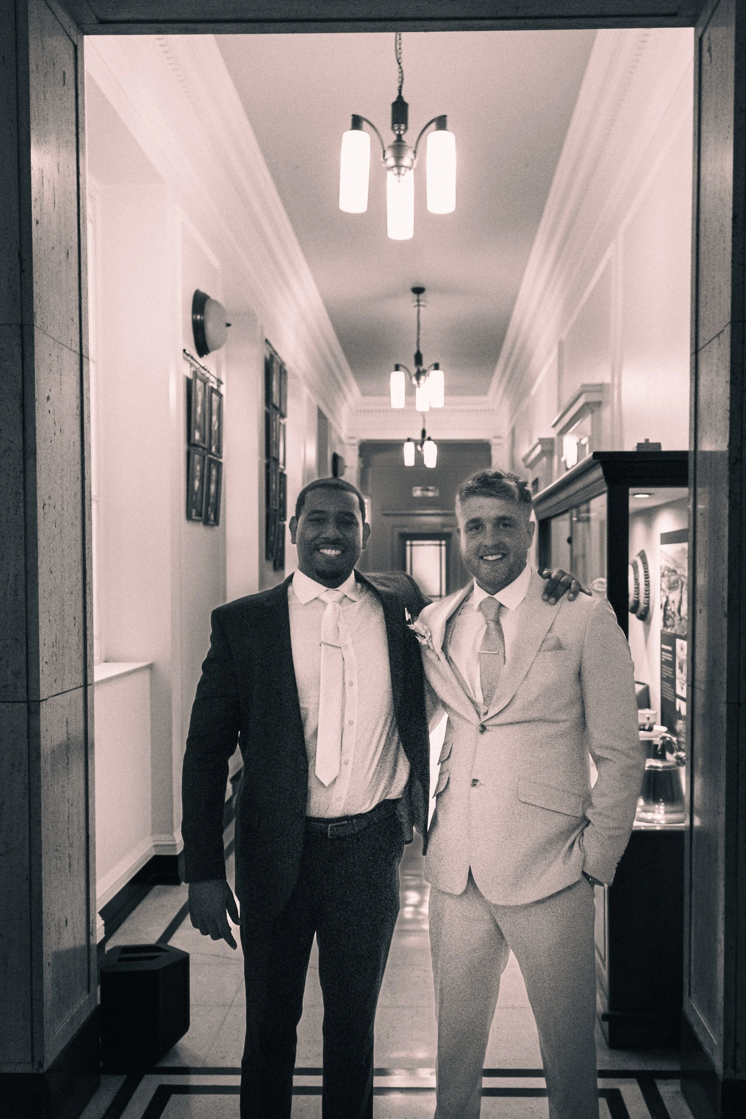 Two men in suits smiling, standing together in hallway with ceiling lights and framed pictures on the wall.