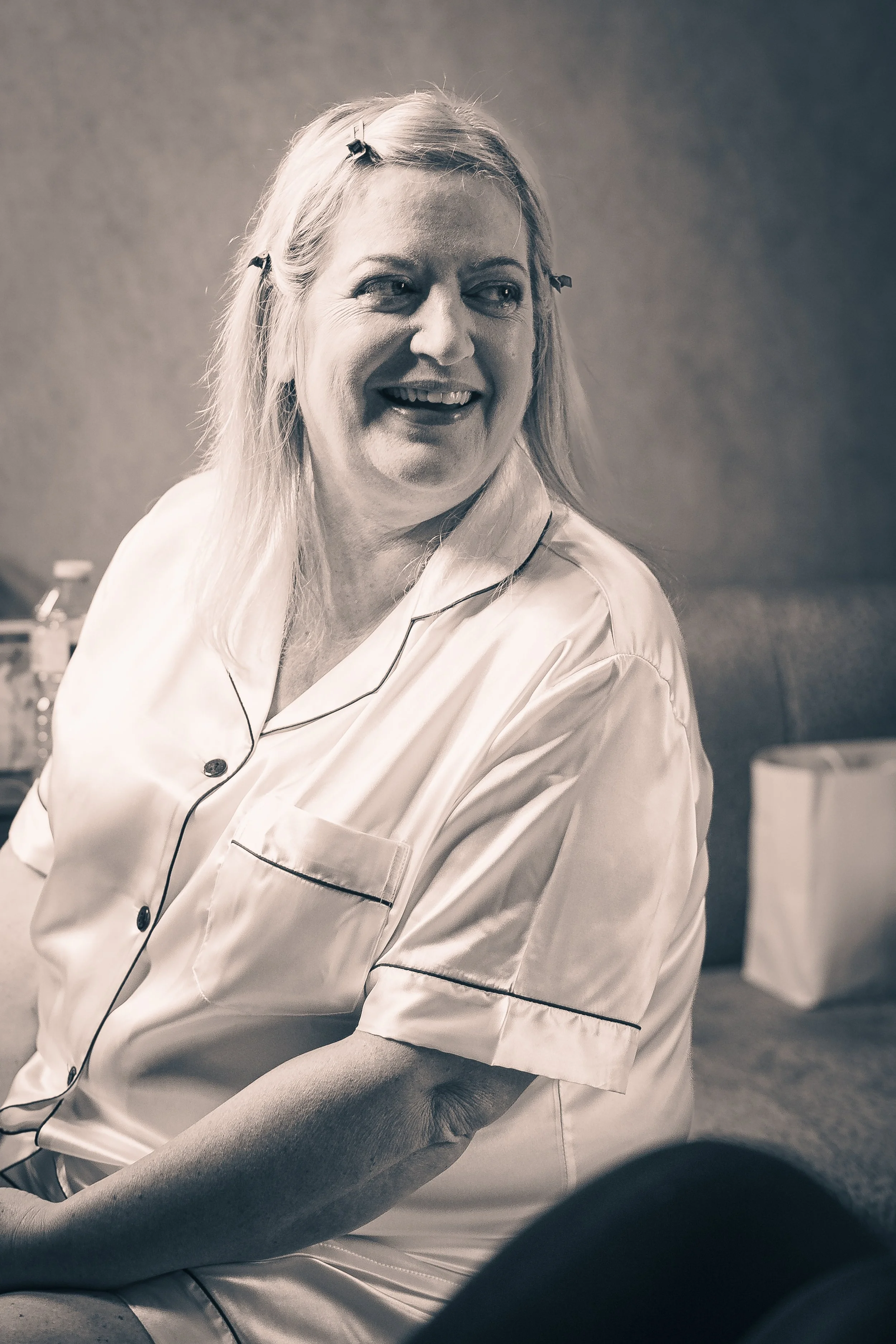 A woman with long blonde hair and hair clips, dressed in pajamas, smiling and sitting on a bed in a room.
