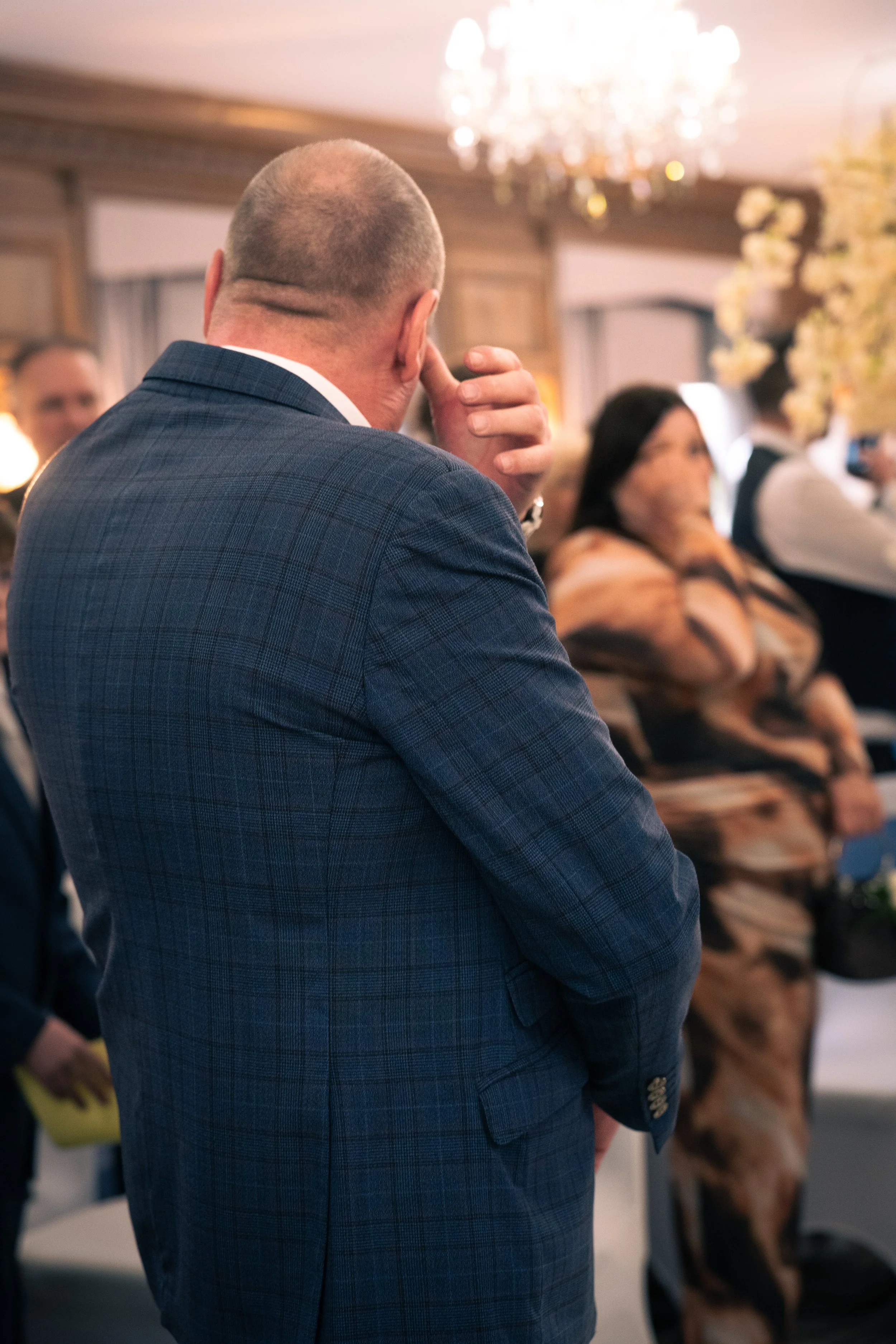 A man with a bald head wearing a blue checkered suit is seen from behind, touching his face with his hand. He is in a formal event with other people, a chandelier hanging from the ceiling, and a floral arrangement in the background.