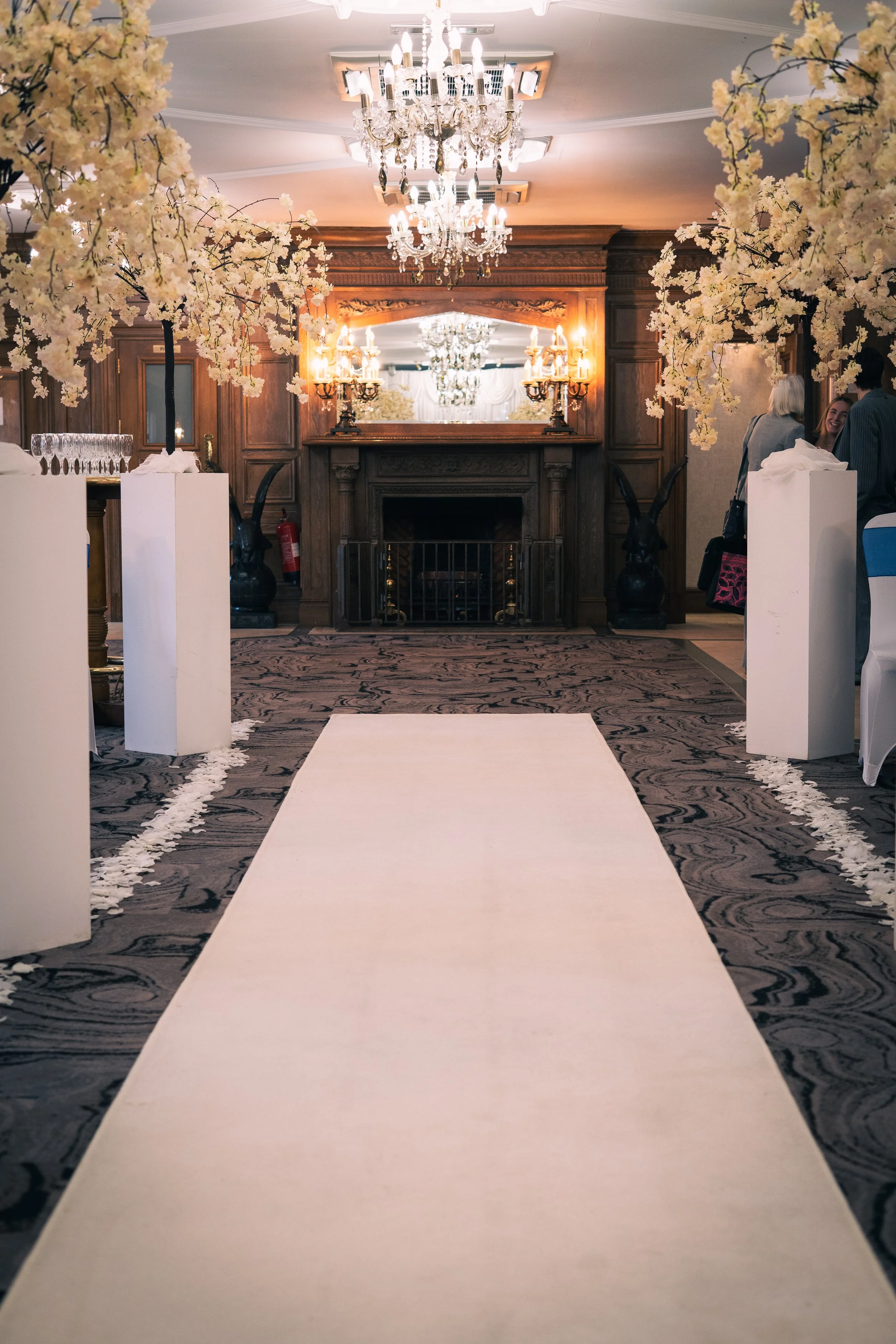 Elegant indoor wedding ceremony setup with a white aisle runner, floral arrangements, chandeliers, and a fireplace