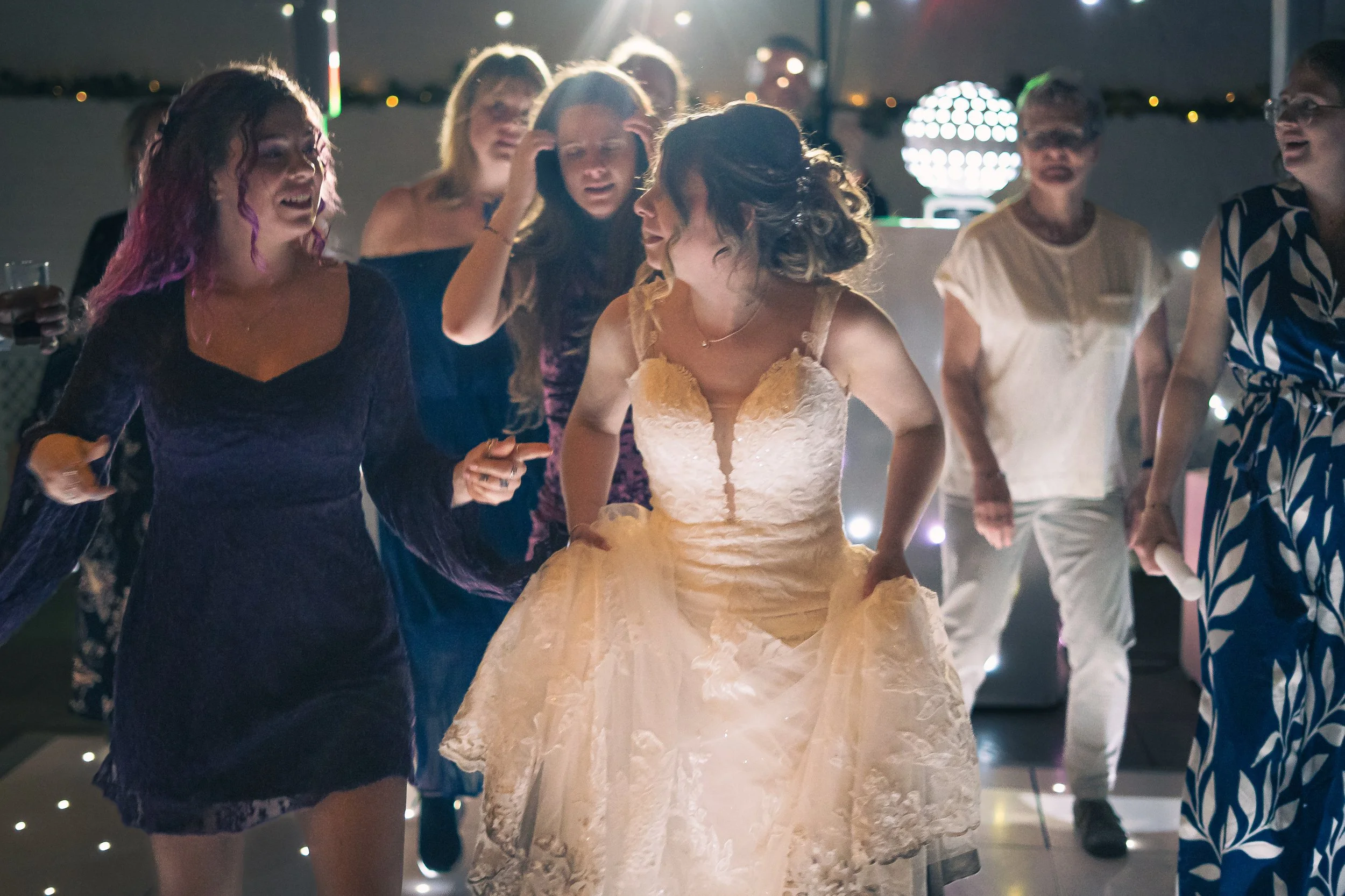 Group of women dancing at a celebration, one is in a wedding dress, at night with decorated lighting in the background.