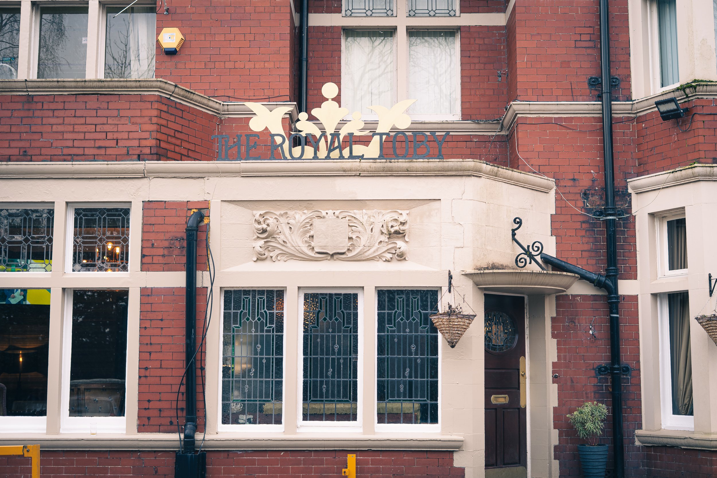 The front exterior of a brick building named 'The Royal Toby,' featuring stained glass windows, a decorative stone crest, hanging baskets, and a black drainpipe.
