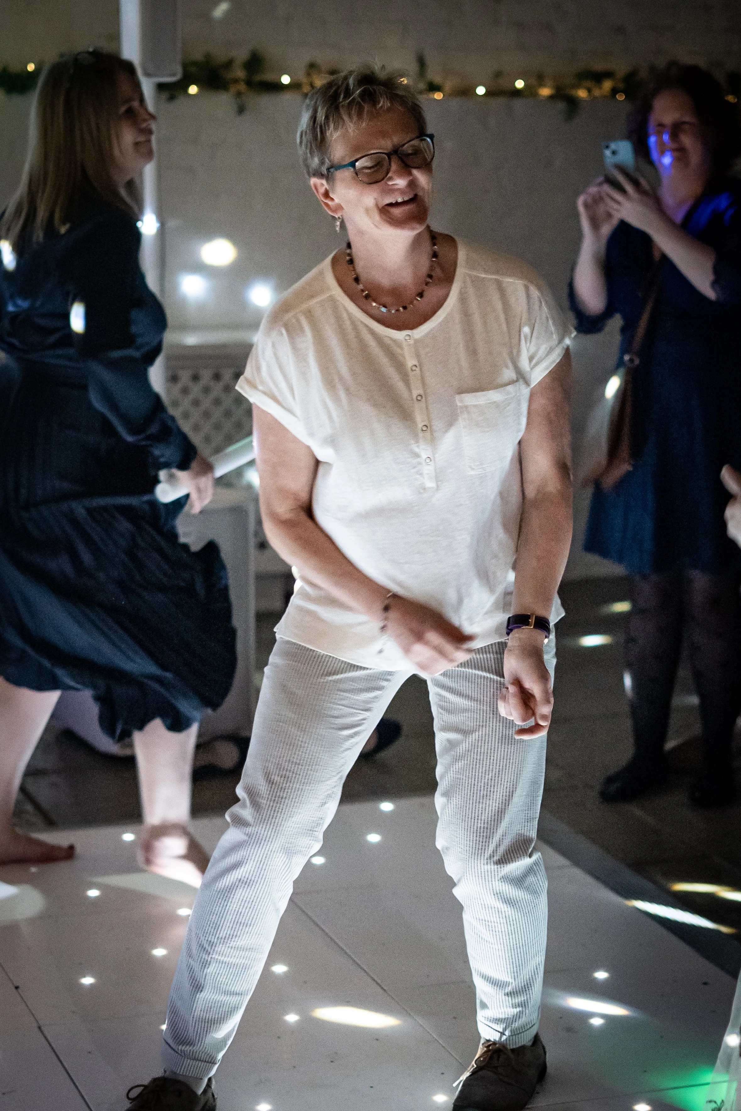 An older woman with glasses and short hair dancing at a party. She is wearing a cream-colored shirt and striped pants, smiling, with other women in the background taking pictures and enjoying the moment.
