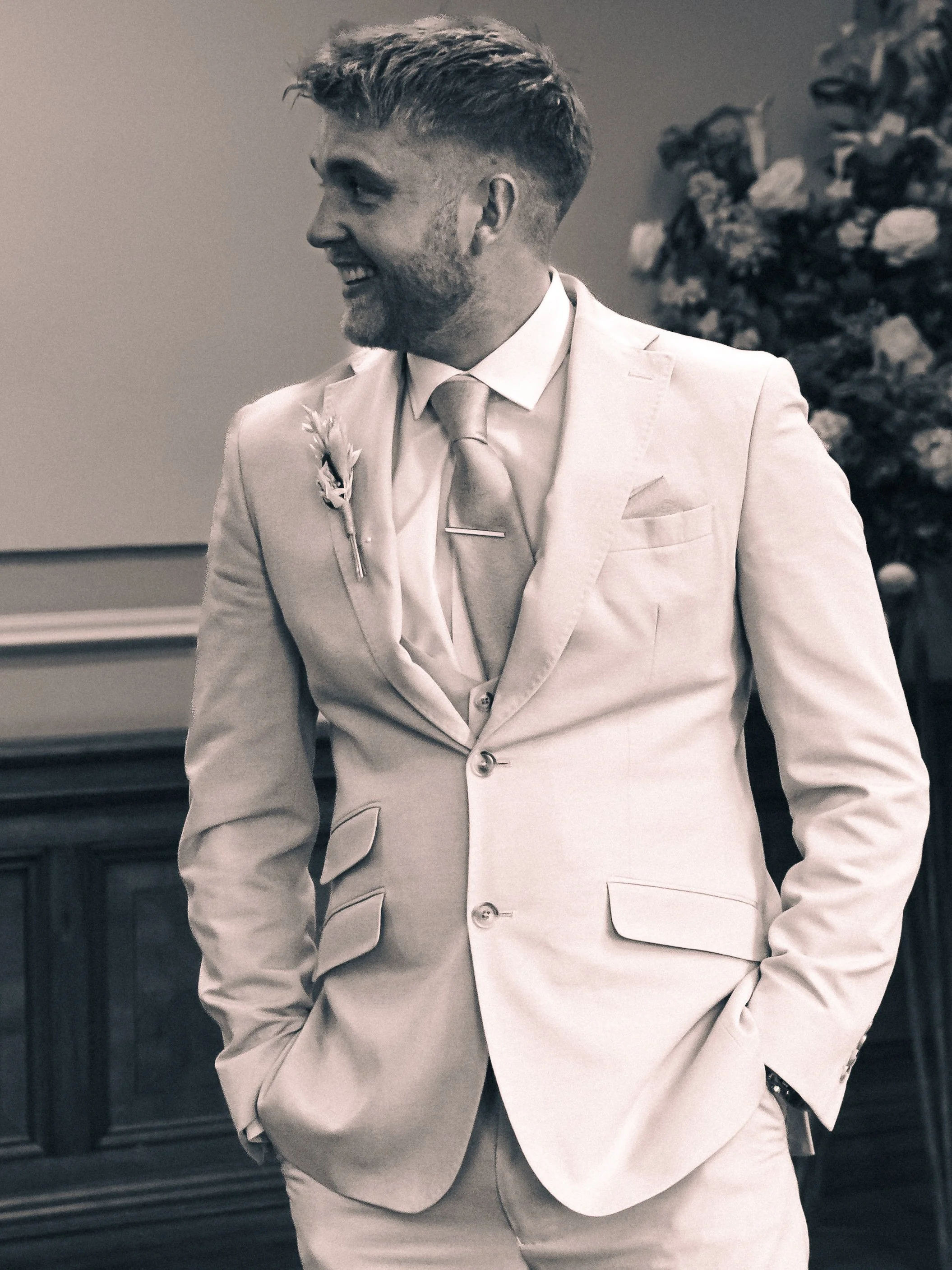 A man in a light-colored suit, white shirt, and tie standing indoors, smiling and looking to the side, with a blurred background featuring a floral arrangement.