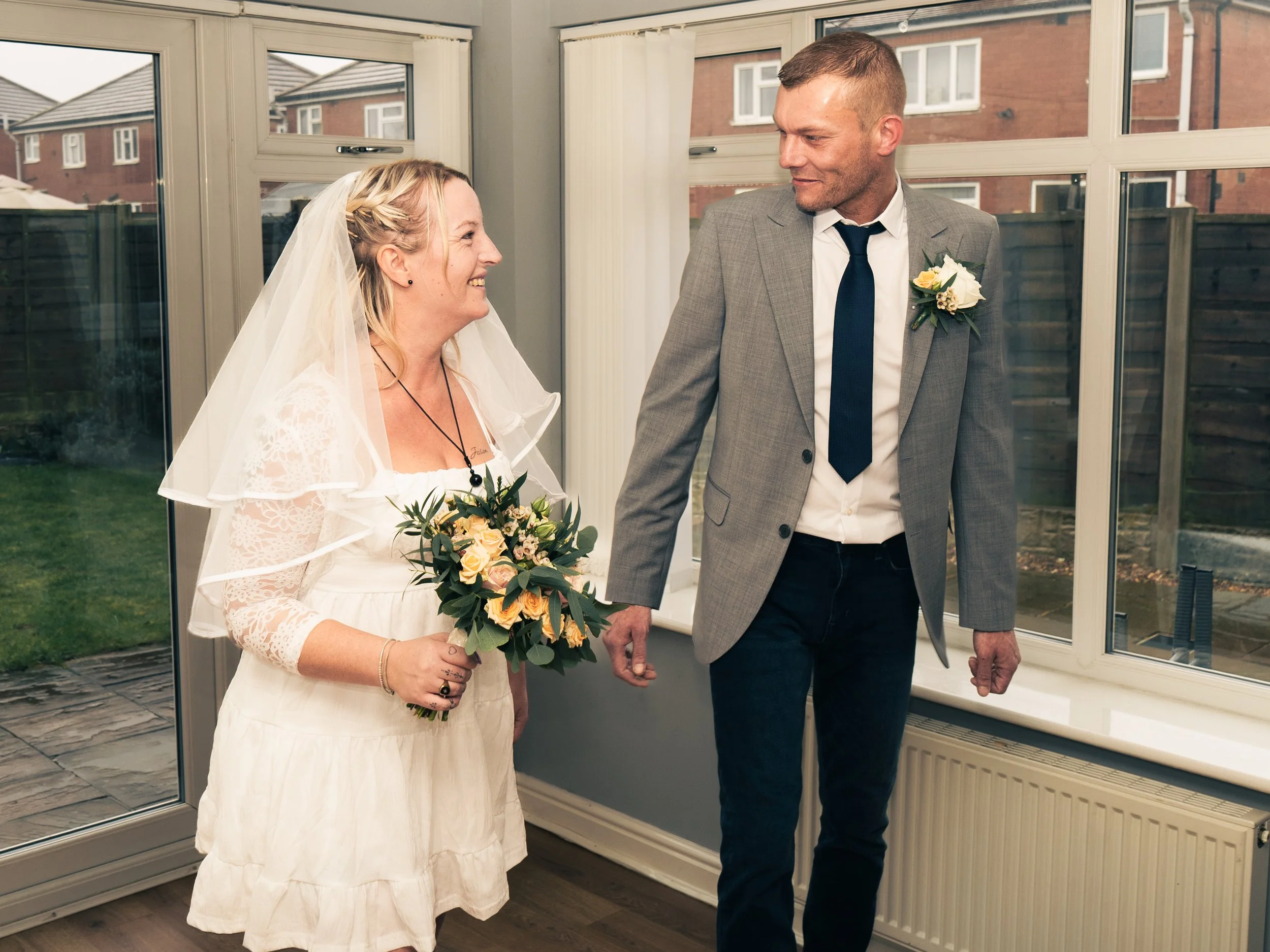 A bride and groom holding hands inside a room with large windows, smiling at each other, during their wedding celebration.