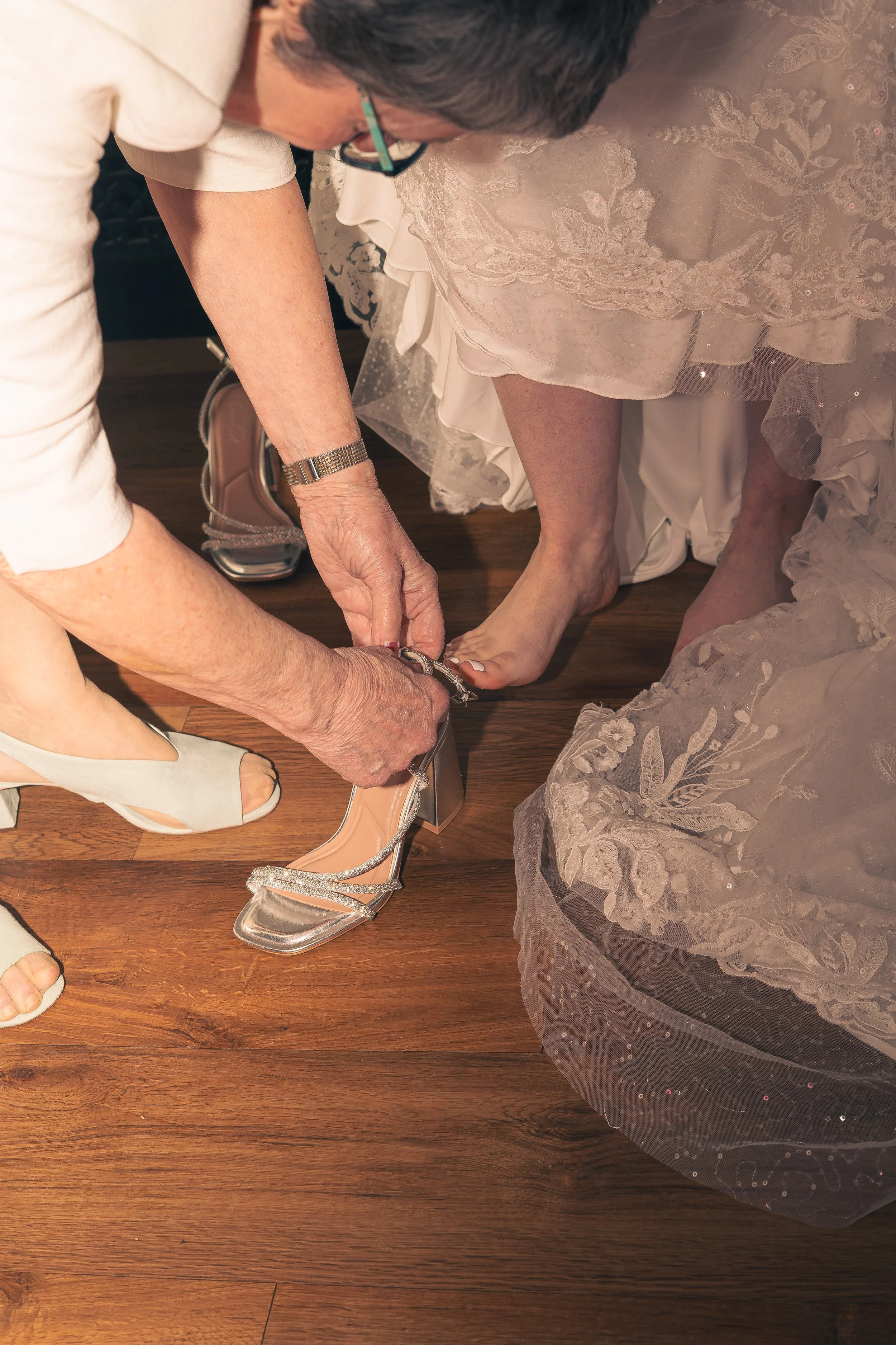 An elderly woman helping a bride put on her wedding shoes on a wooden floor. The bride's dress is light-colored with lace details and she is barefoot, while the woman is wearing light-colored shoes.