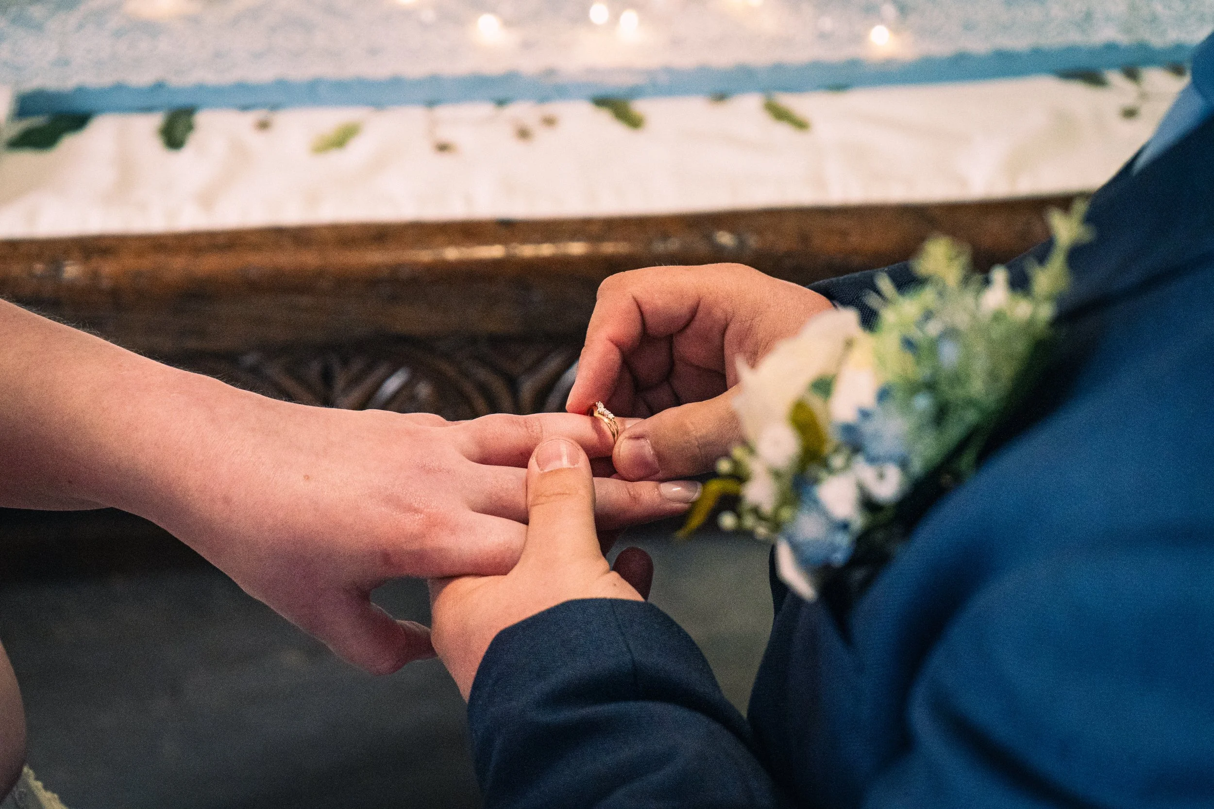 Close-up of a wedding ceremony where one person is placing a ring on the other's finger, with a blurred flower arrangement in the foreground.