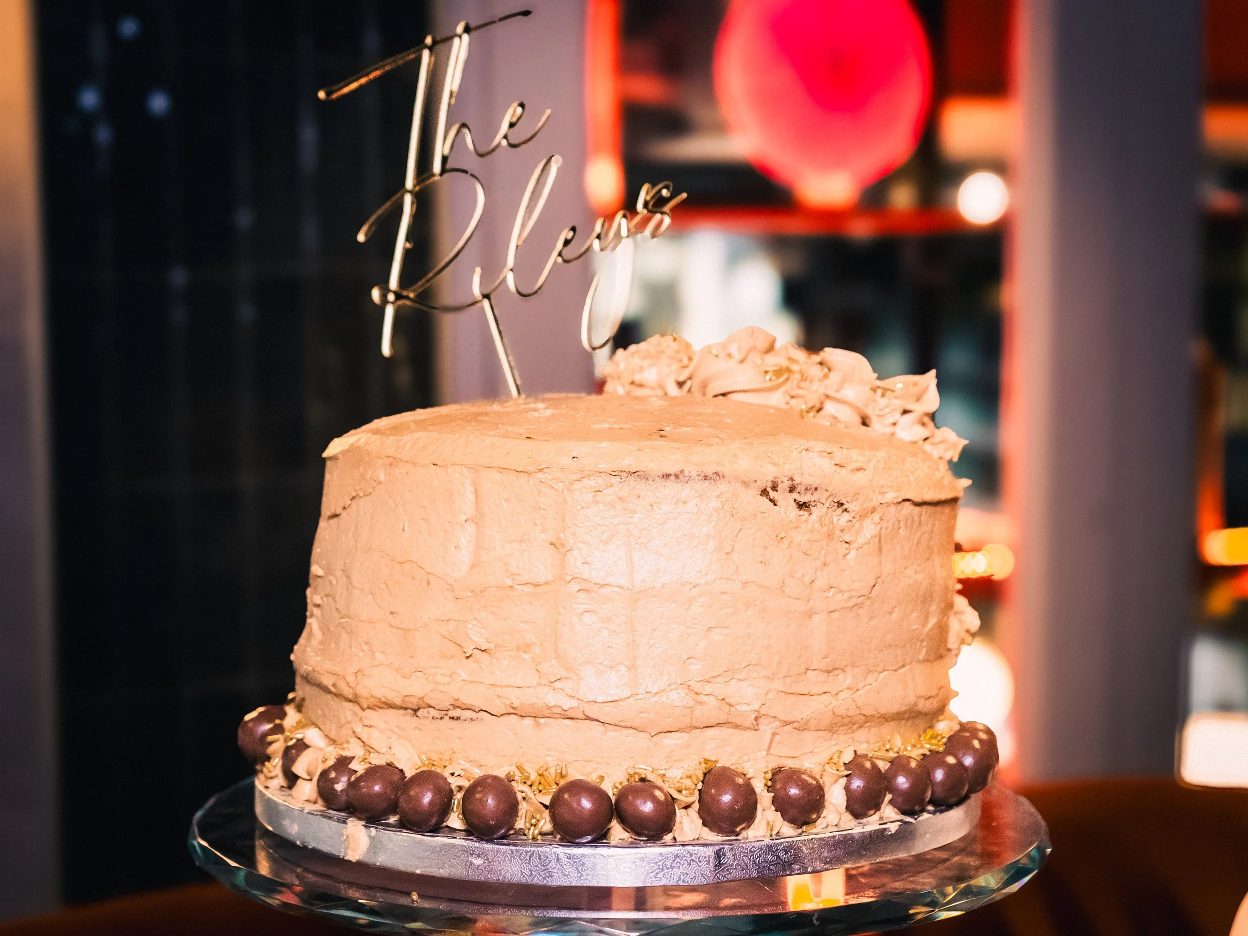 A round birthday cake with beige frosting, decorated with dark chocolate spheres and an ornate border, topped with a cake topper that reads, 'The Rebel'.