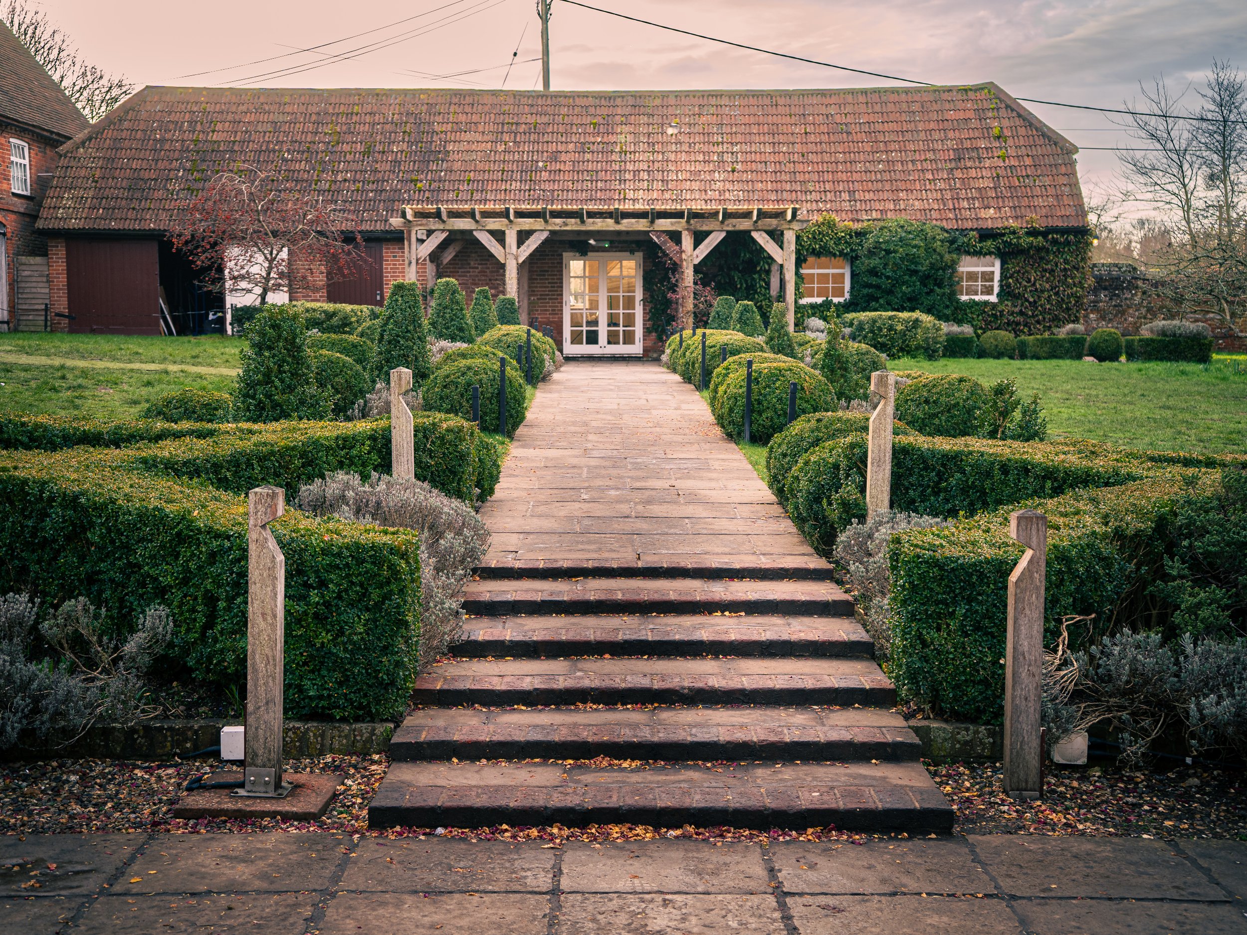 A brick pathway with steps leading up to a house entrance, surrounded by well-maintained bushes and greenery, with a garden and a barn-like structure in the background.