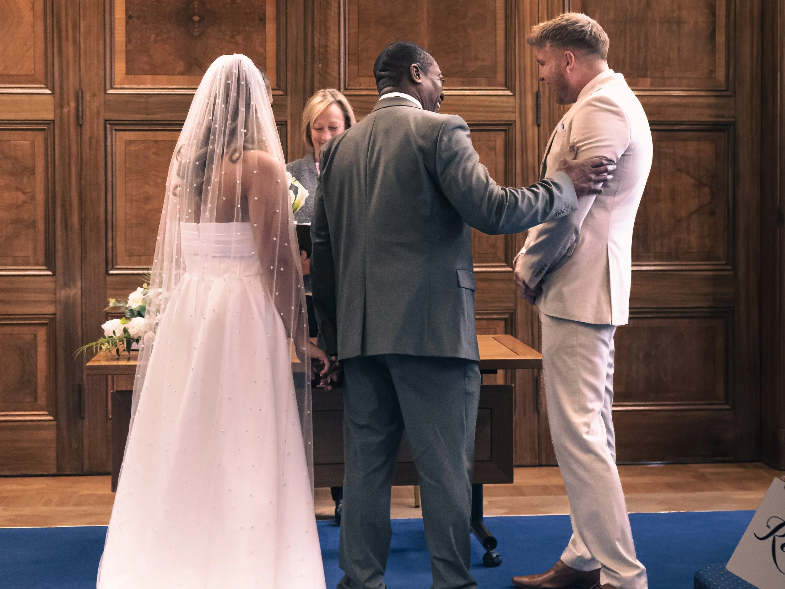 A wedding ceremony with a bride, groom, and officiant standing in front of a wooden wall. The bride is in a white wedding dress and veil, the groom is in a white suit, and the officiant is in a gray suit.