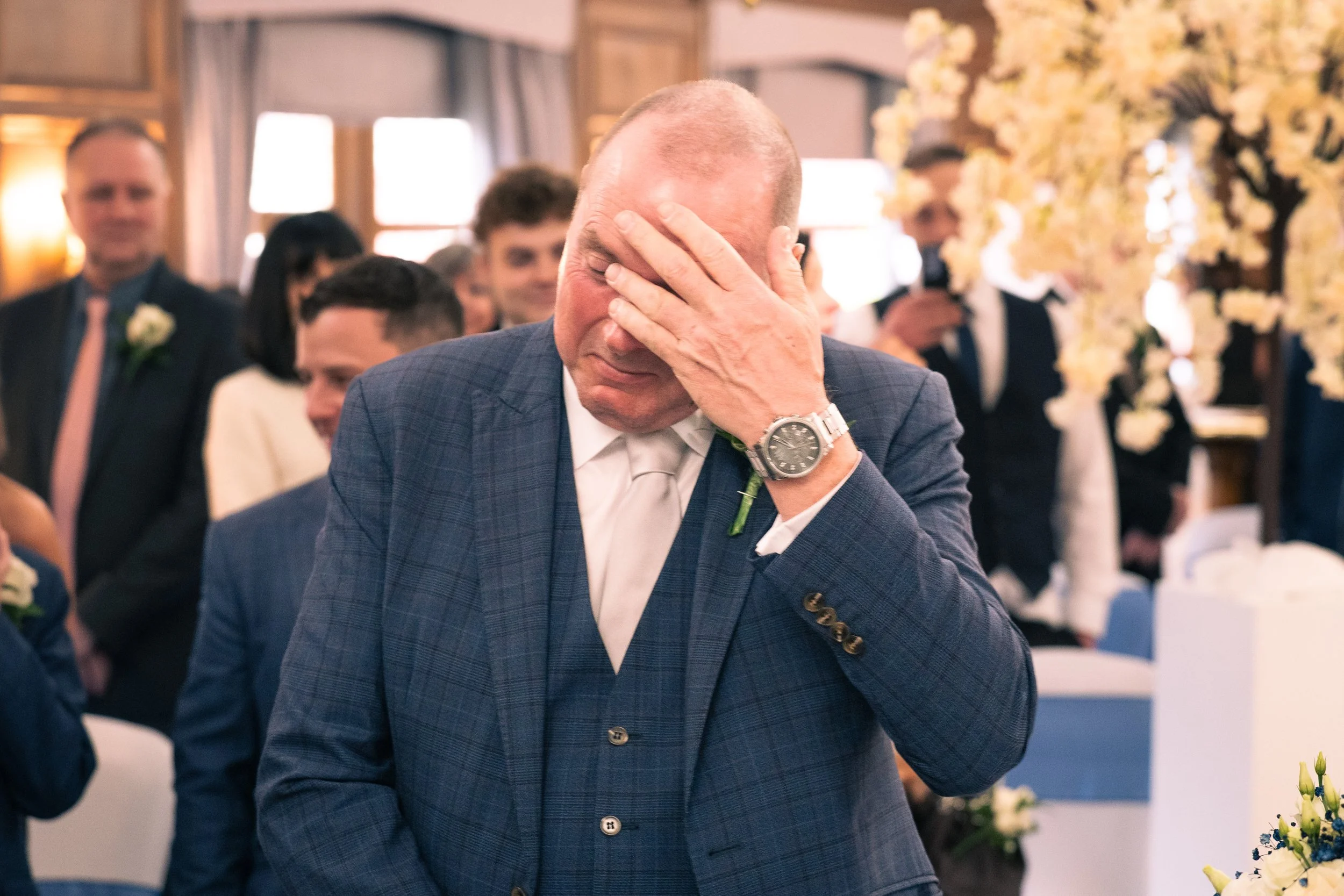 A man in a blue plaid suit and white tie with a boutonniere on his lapel, holding his face with his hand, standing in a decorated room with other people in the background, appearing emotional.