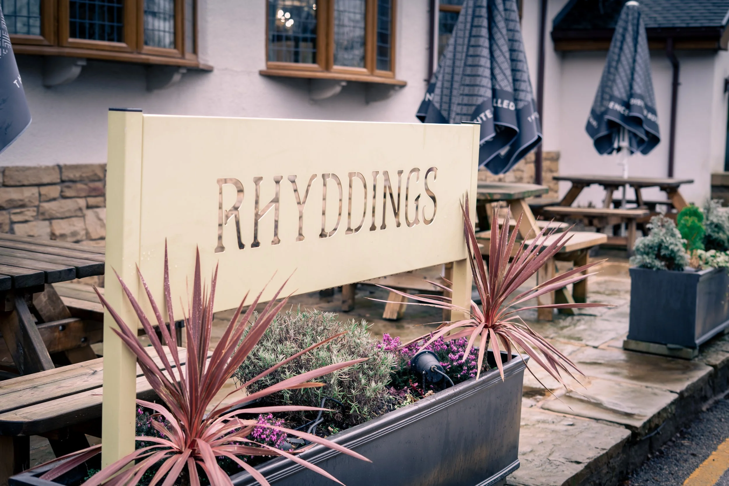 Wooden outdoor seating area with tables and benches, potted plants, and a sign that reads 'RHYDDINGS' in front of a building with windows and umbrellas.