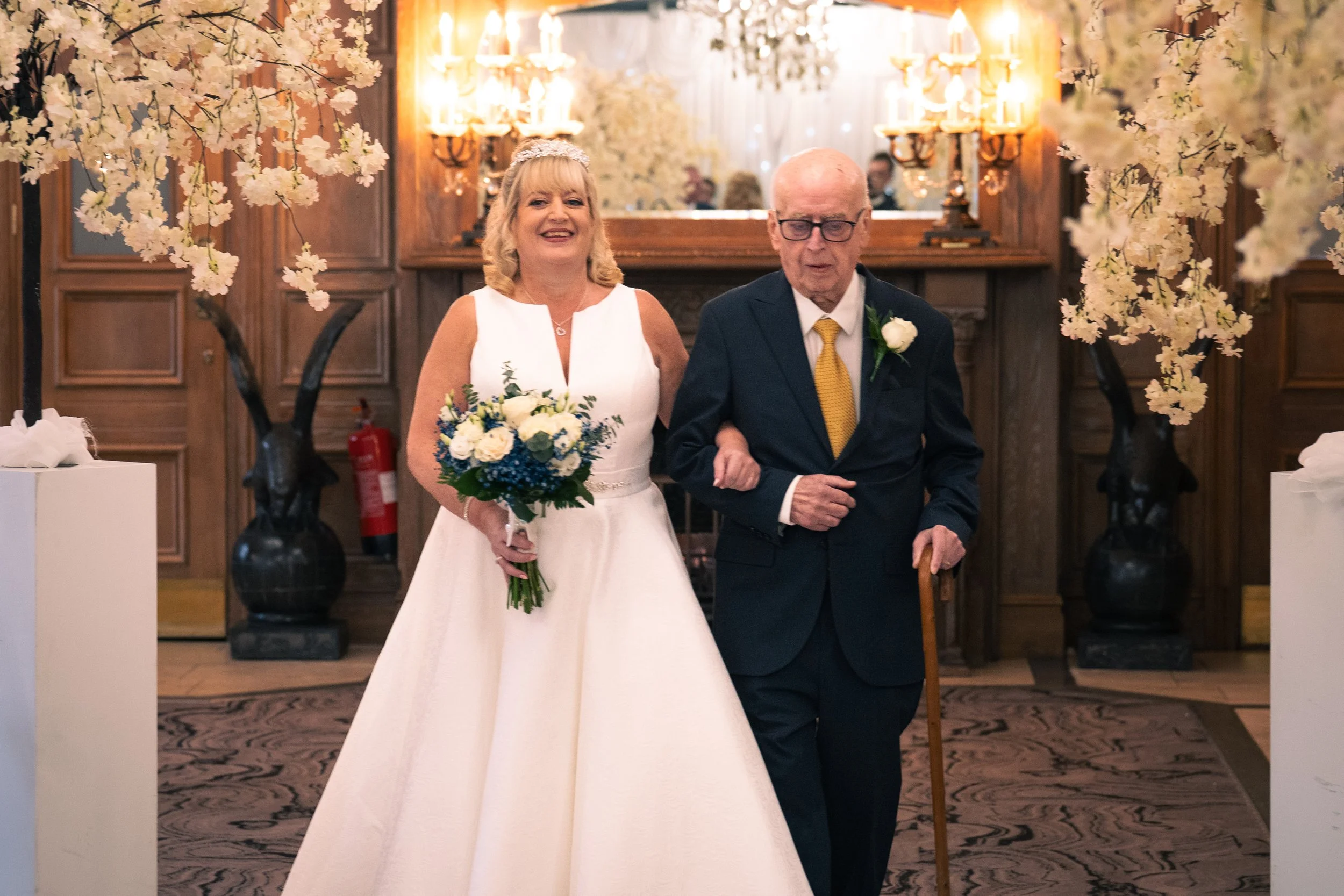 An elderly man with glasses and a cane walking down the aisle with a smiling woman in a white wedding dress, holding a bouquet, in a decorated wedding venue with floral arrangements and warm lighting.