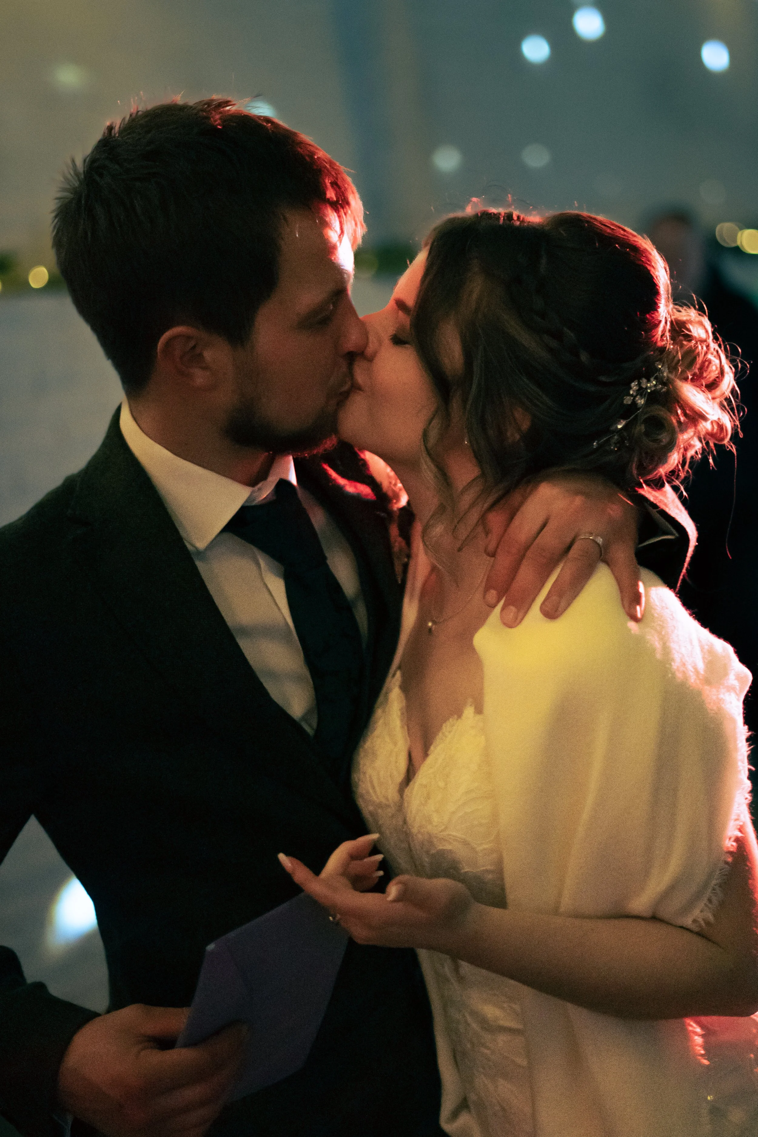 A man and woman share a kiss at a wedding reception, with a dark background and soft lighting.