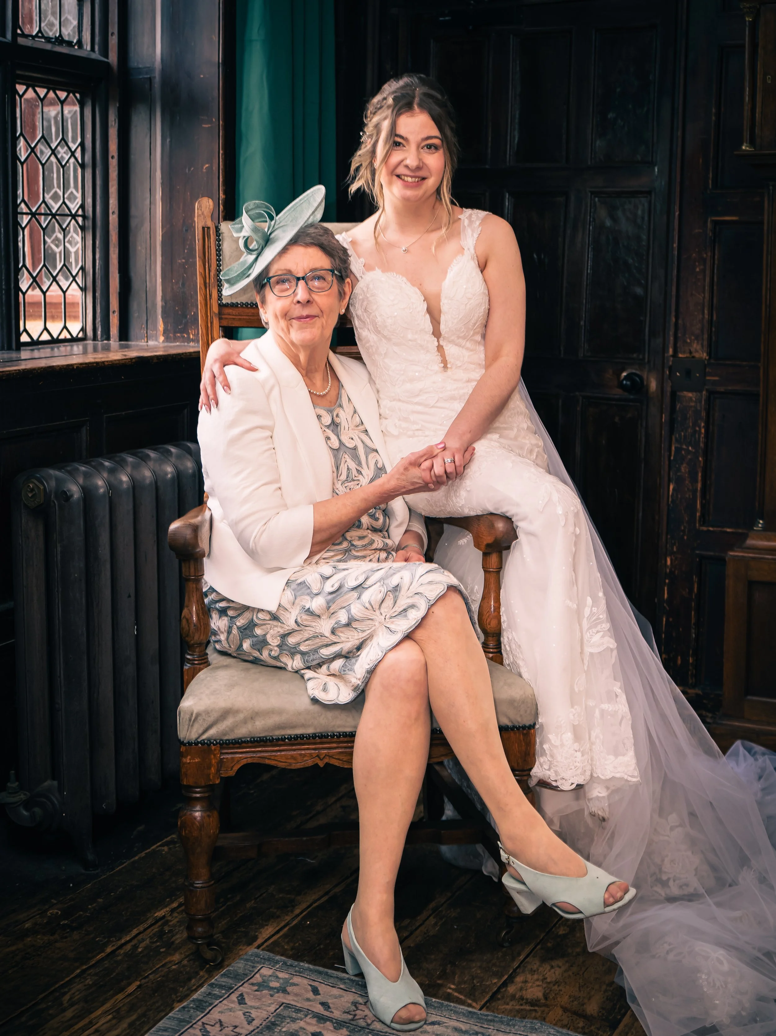 A young woman in a wedding dress sitting on a woman's lap, both smiling, in a vintage-style room with dark wood paneling and stained glass windows.