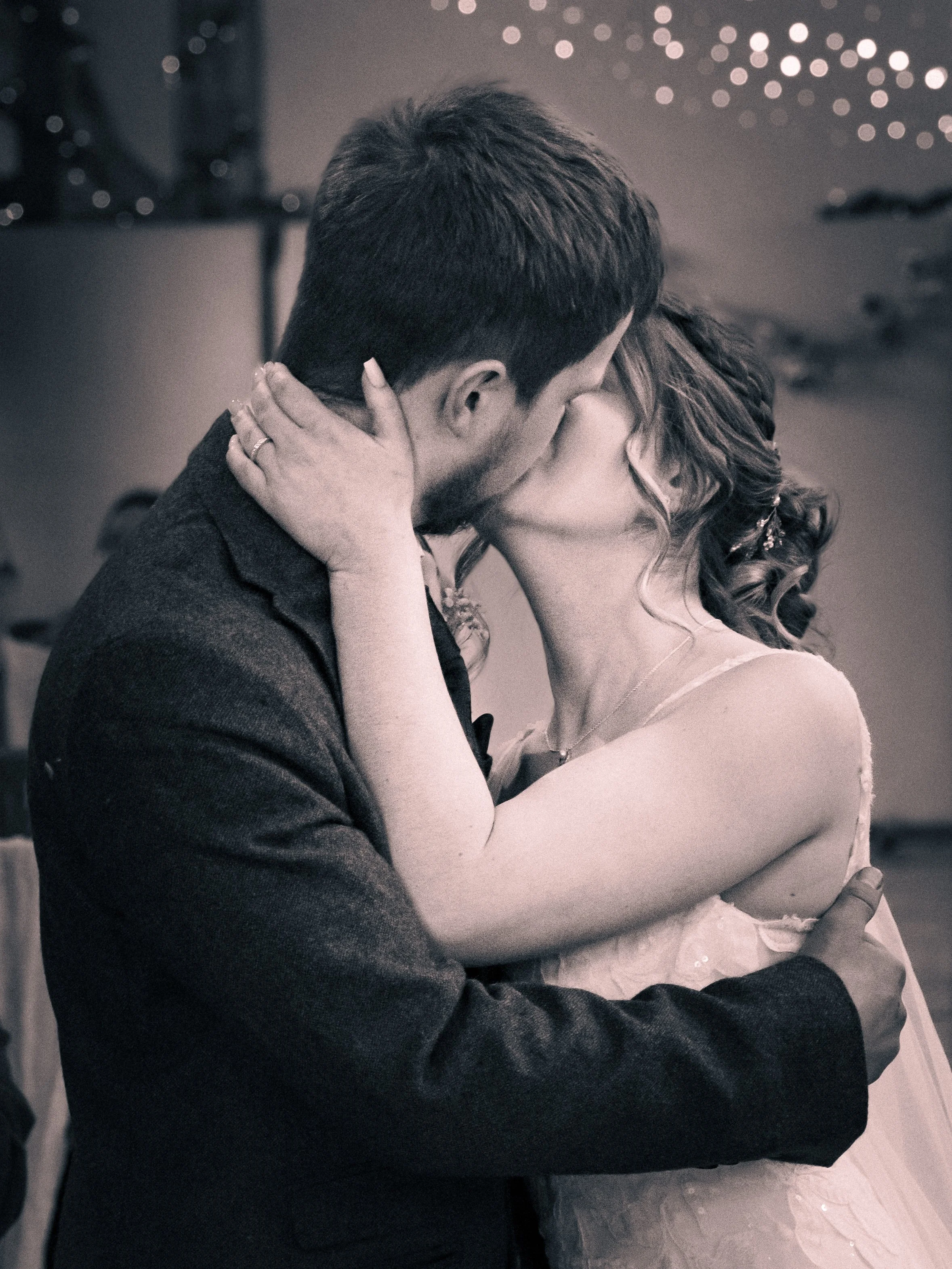 A couple kissing passionately at a wedding or celebration, with festive lights in the background.