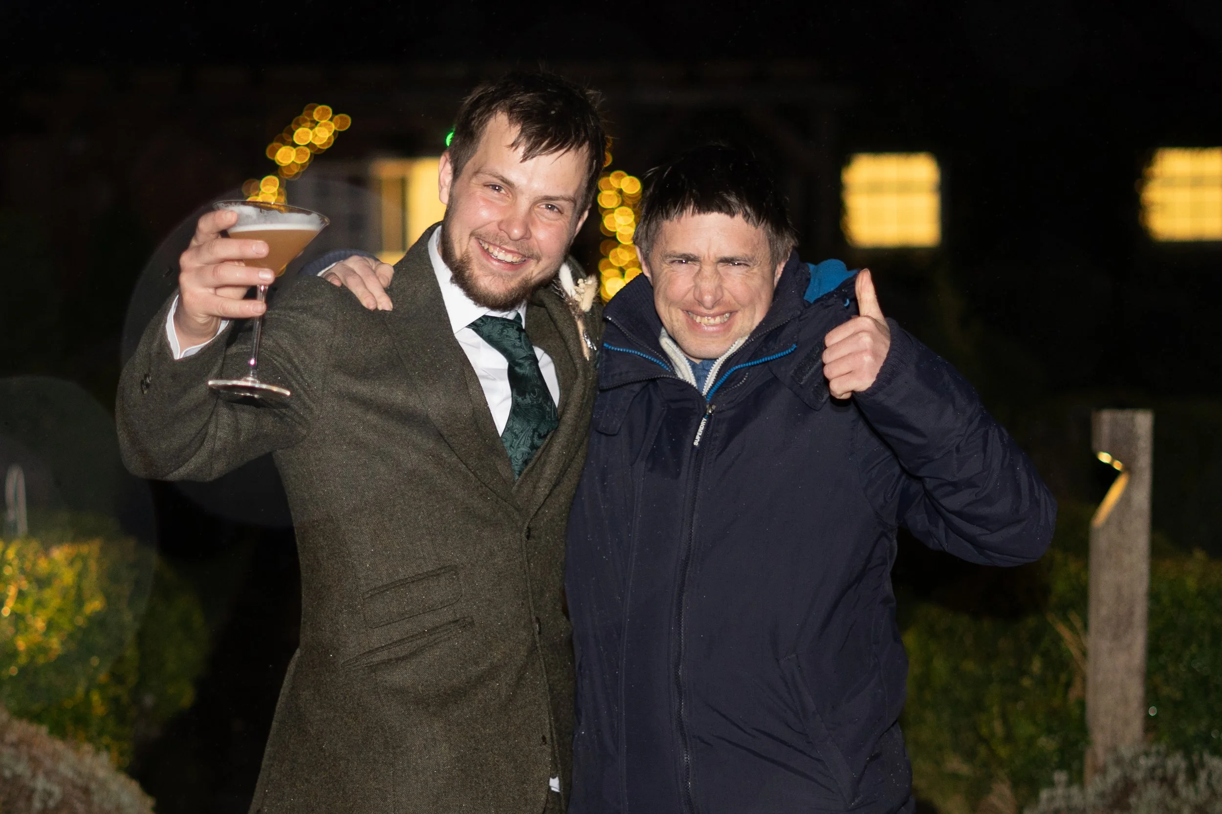 Two men standing outside at night, smiling and celebrating; one holds a cocktail glass, the other gives a thumbs-up.