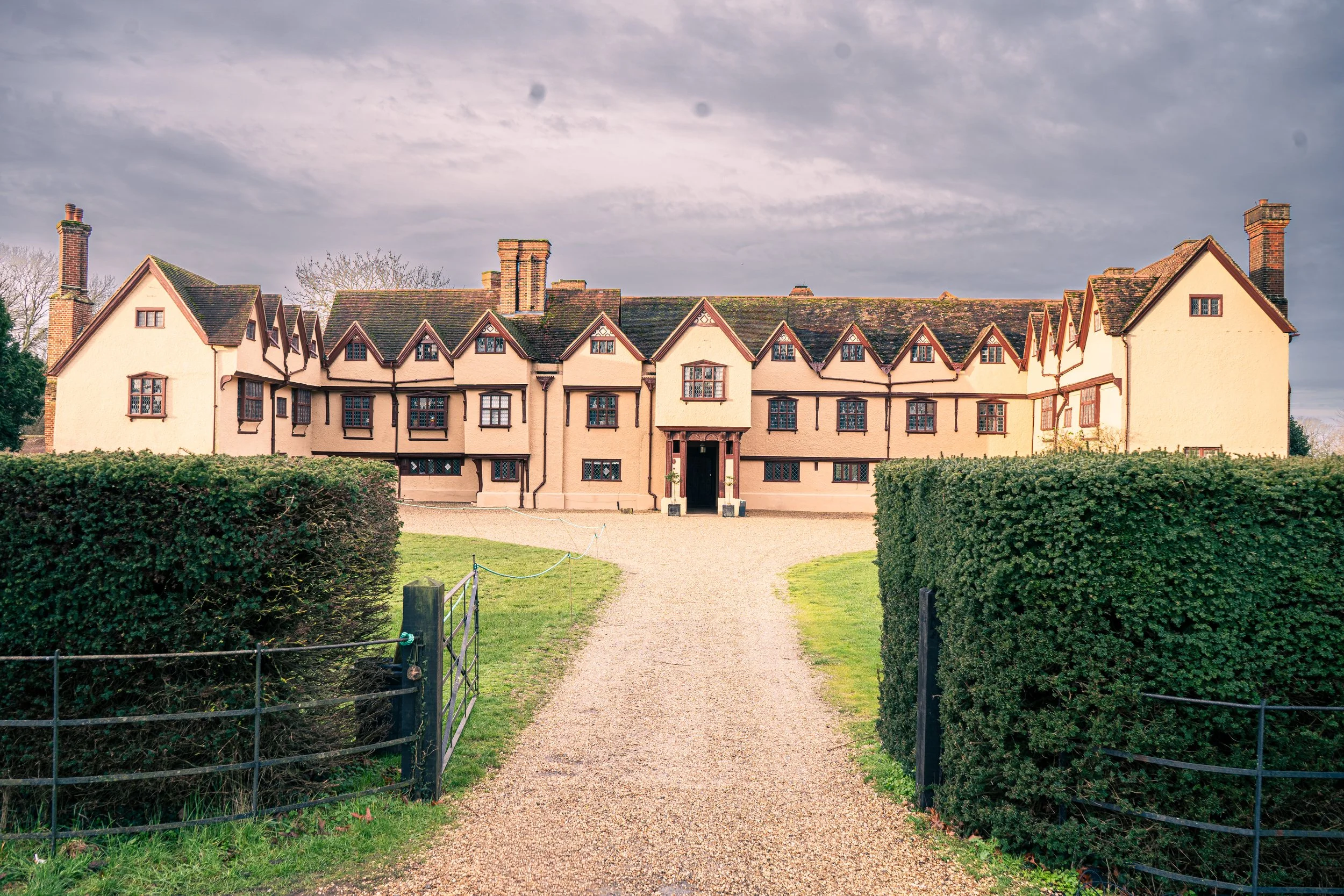 A historic multi-story house with a cream-colored exterior, multiple gabled roofs, and several chimneys, surrounded by a gravel pathway and trimmed hedges, under a cloudy sky.