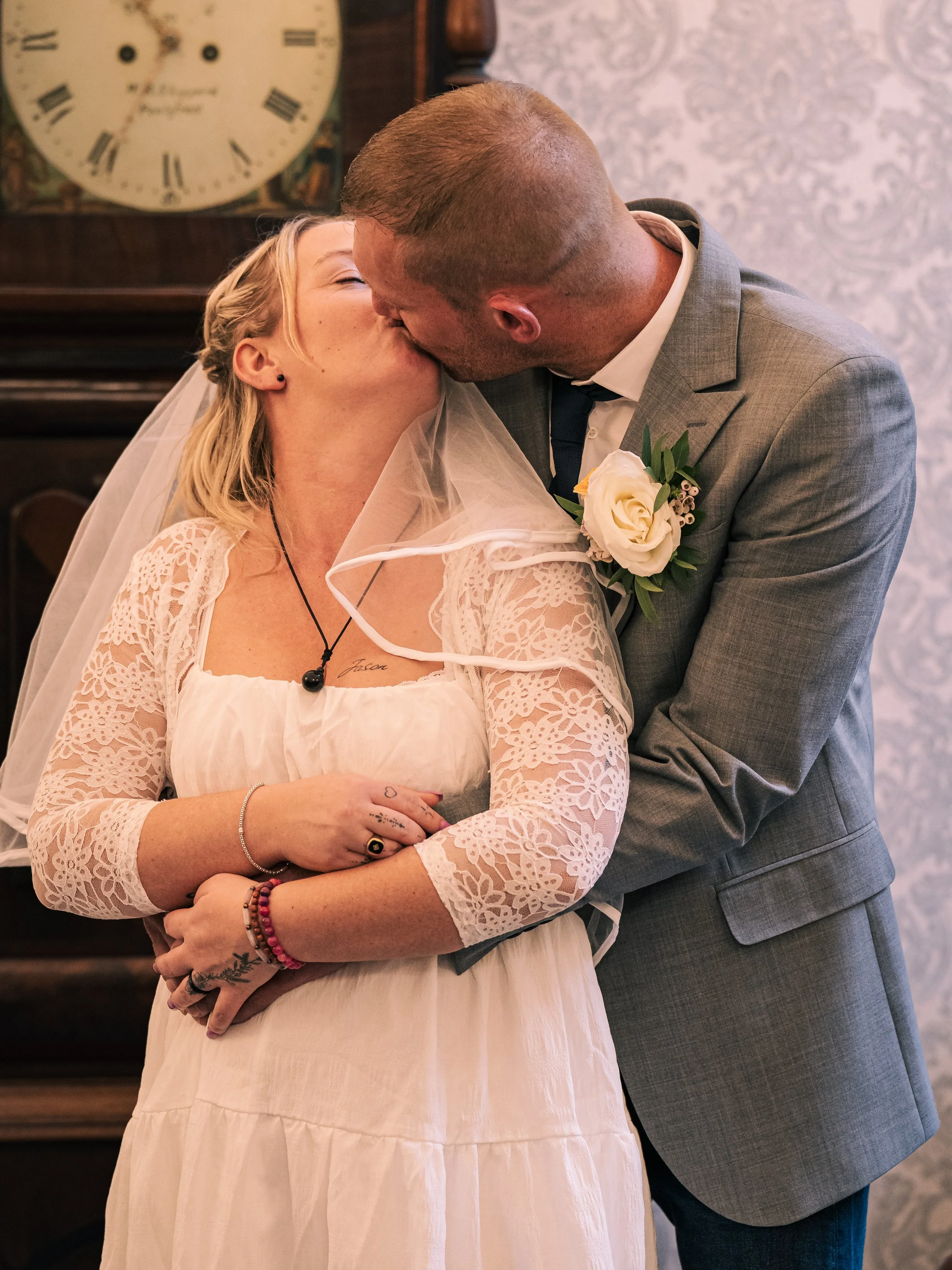 A couple kissing at their wedding ceremony, with the bride wearing a white lace dress and veil, and the groom in a light gray suit with a white boutonniere.