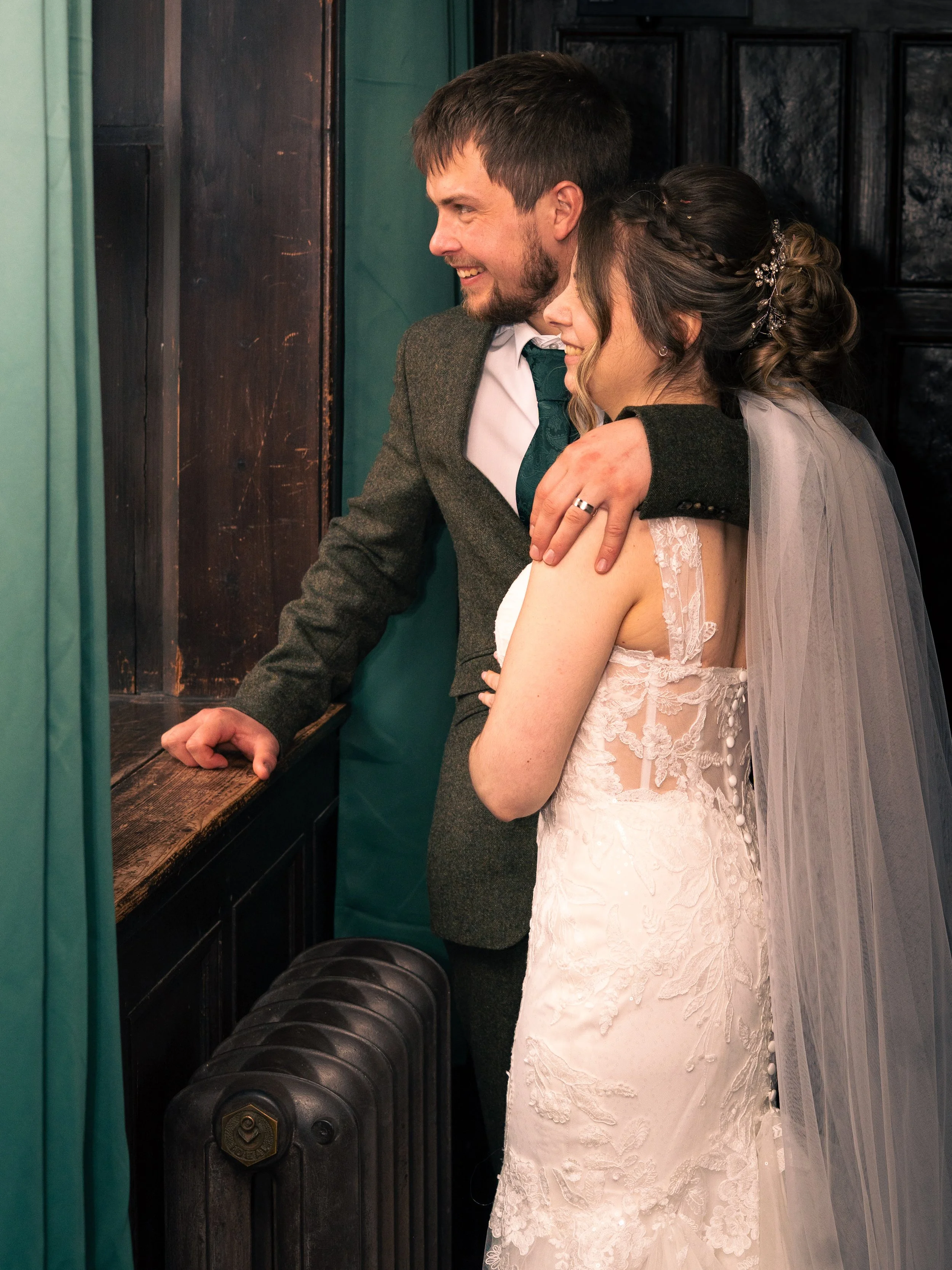 A newlywed couple hugging and smiling, with the bride in a lace wedding gown and veil and the groom in a suit, standing inside a booth or a small room with dark wood paneling.