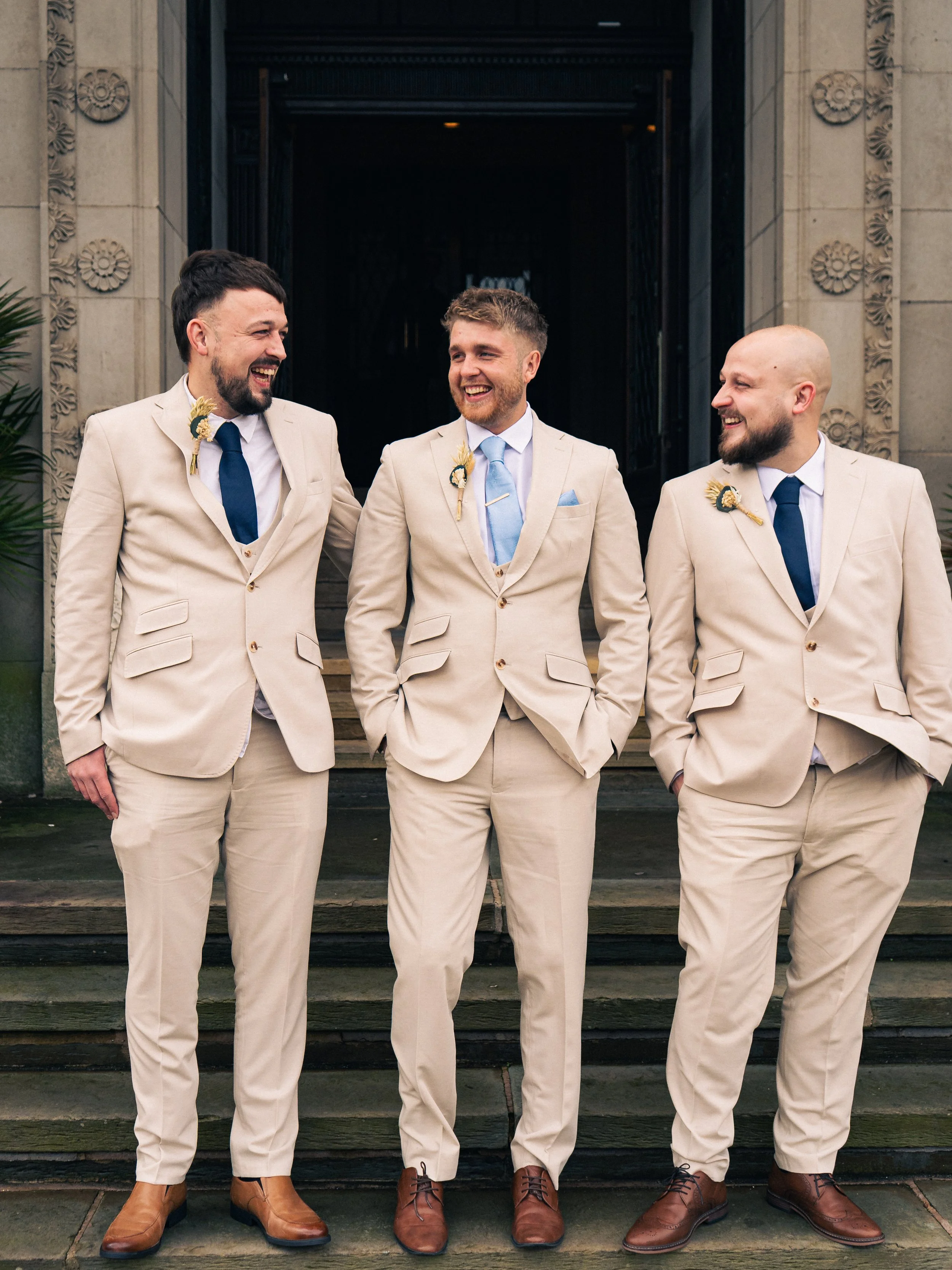 Three men in beige suits and blue ties standing on steps outside a building, smiling and walking together.