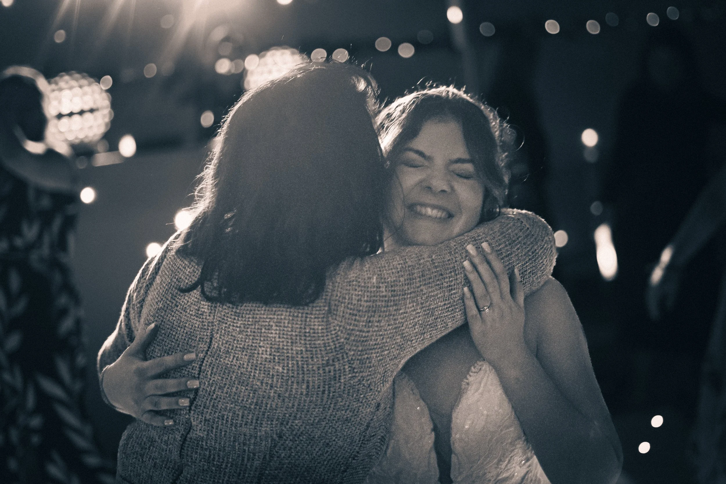 Two women hugging and smiling joyfully at an indoor event with blurred background lights.