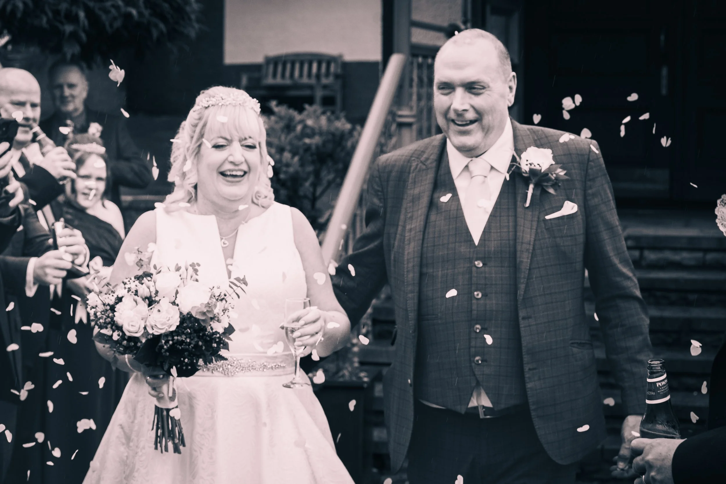 A bride and groom celebrating their wedding, smiling and holding glasses, surrounded by guests throwing confetti.