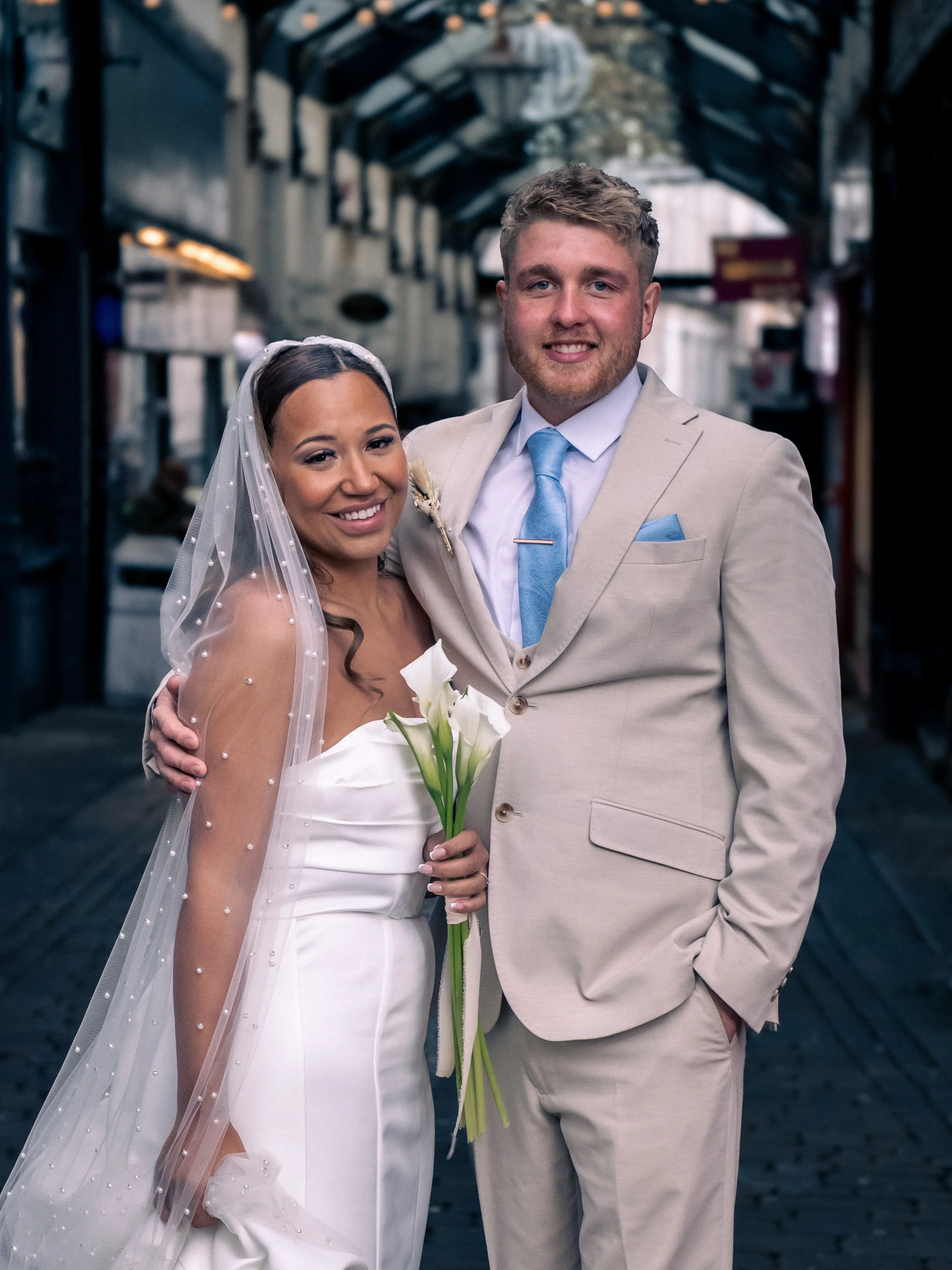 A newlywed couple on their wedding day, standing in a covered outdoor market or walkway. The bride is wearing a white wedding gown with a veil, holding a bouquet of white calla lilies. The groom is dressed in a light beige suit with a blue tie and po