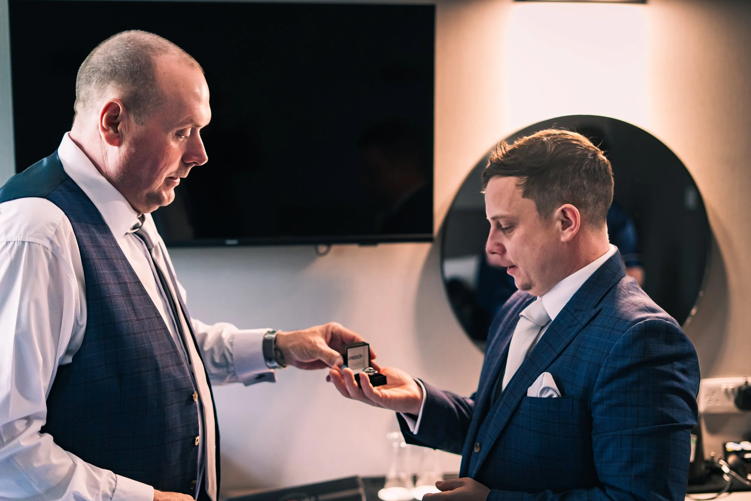 A man in a navy plaid suit receives a ring box from another man in a white shirt and navy plaid vest in a modern room with a large TV and round mirror.