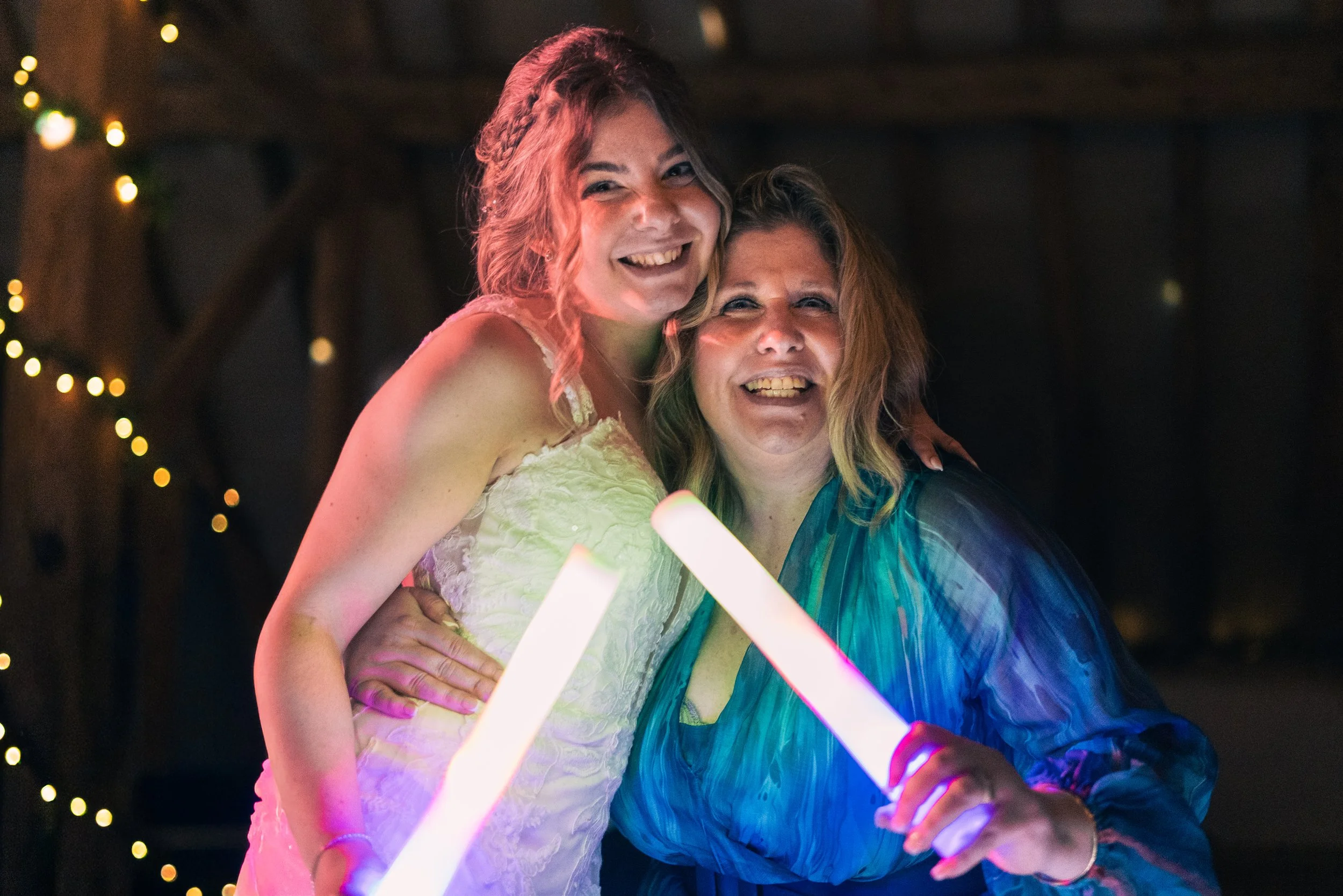Two women smiling and posing for a photo at a celebration, one wearing a white dress and the other wearing a colorful blouse, holding glowing sticks, with string lights in the background.