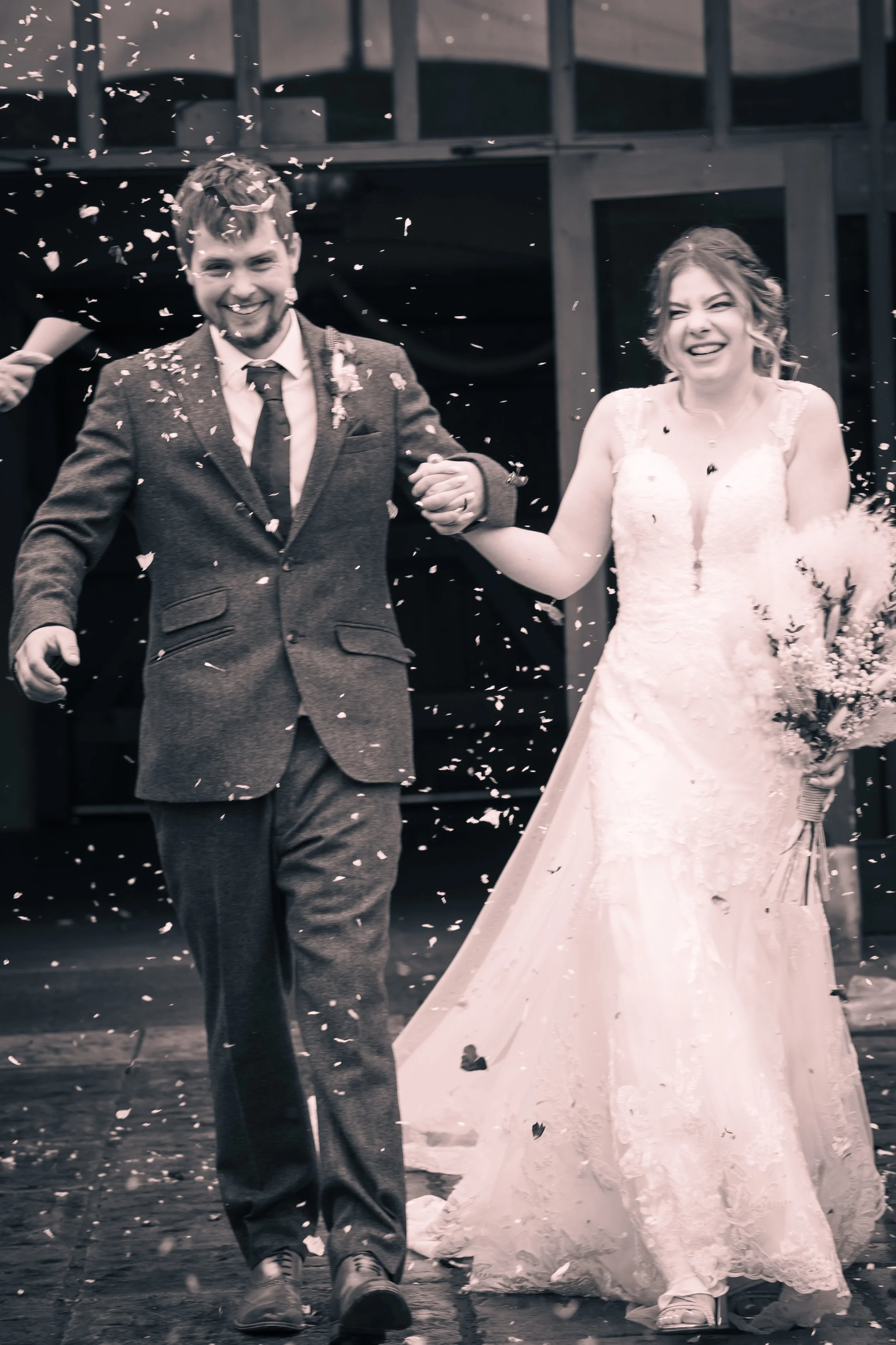 A newlywed couple walking outside, smiling, as confetti is thrown in the air, with the bride holding a bouquet of flowers and wearing a wedding dress, and the groom in a suit.