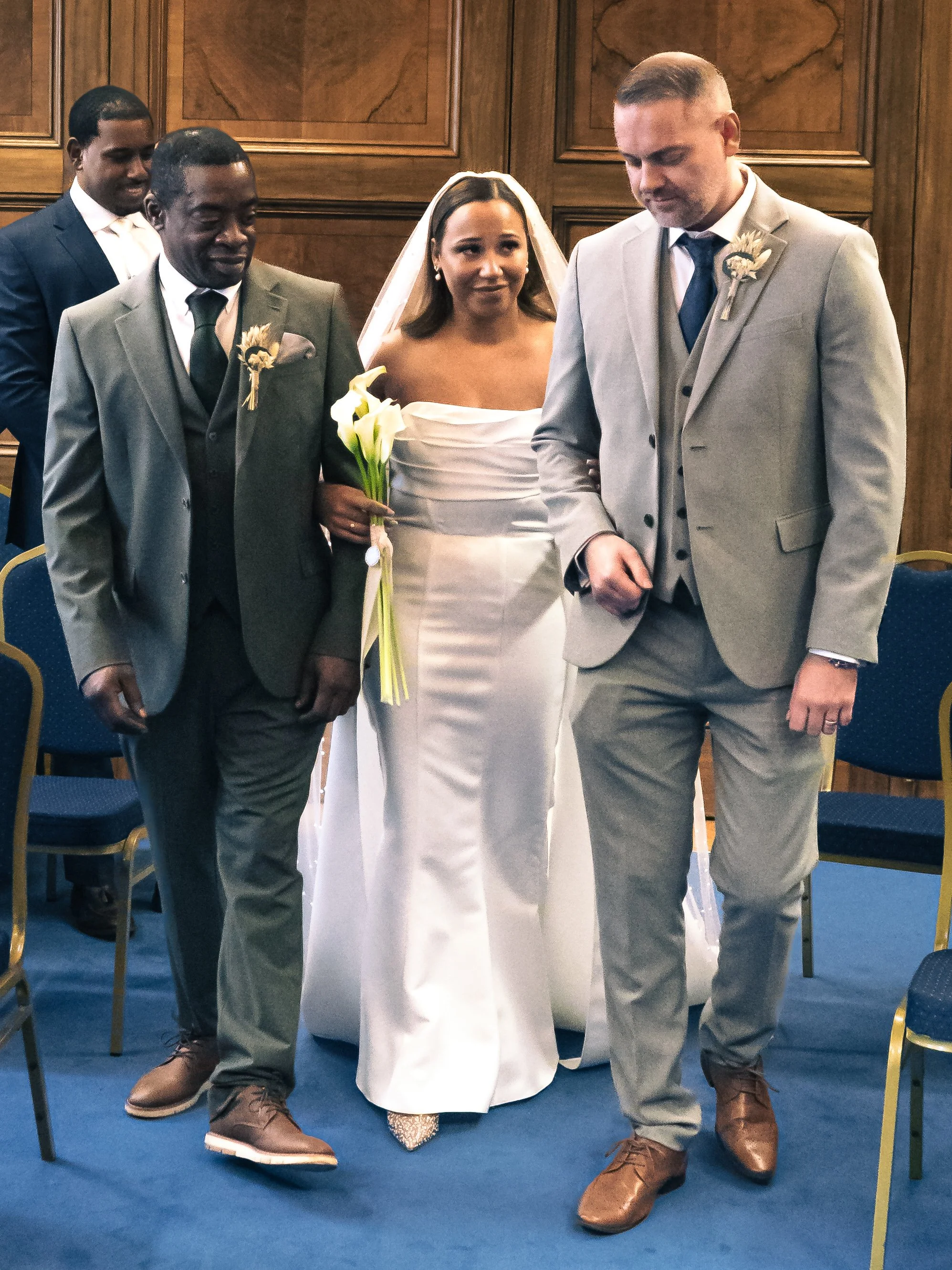 A wedding ceremony with a bride in a white dress holding a bouquet and two men in suits standing next to her, in a room with wood-paneled walls.