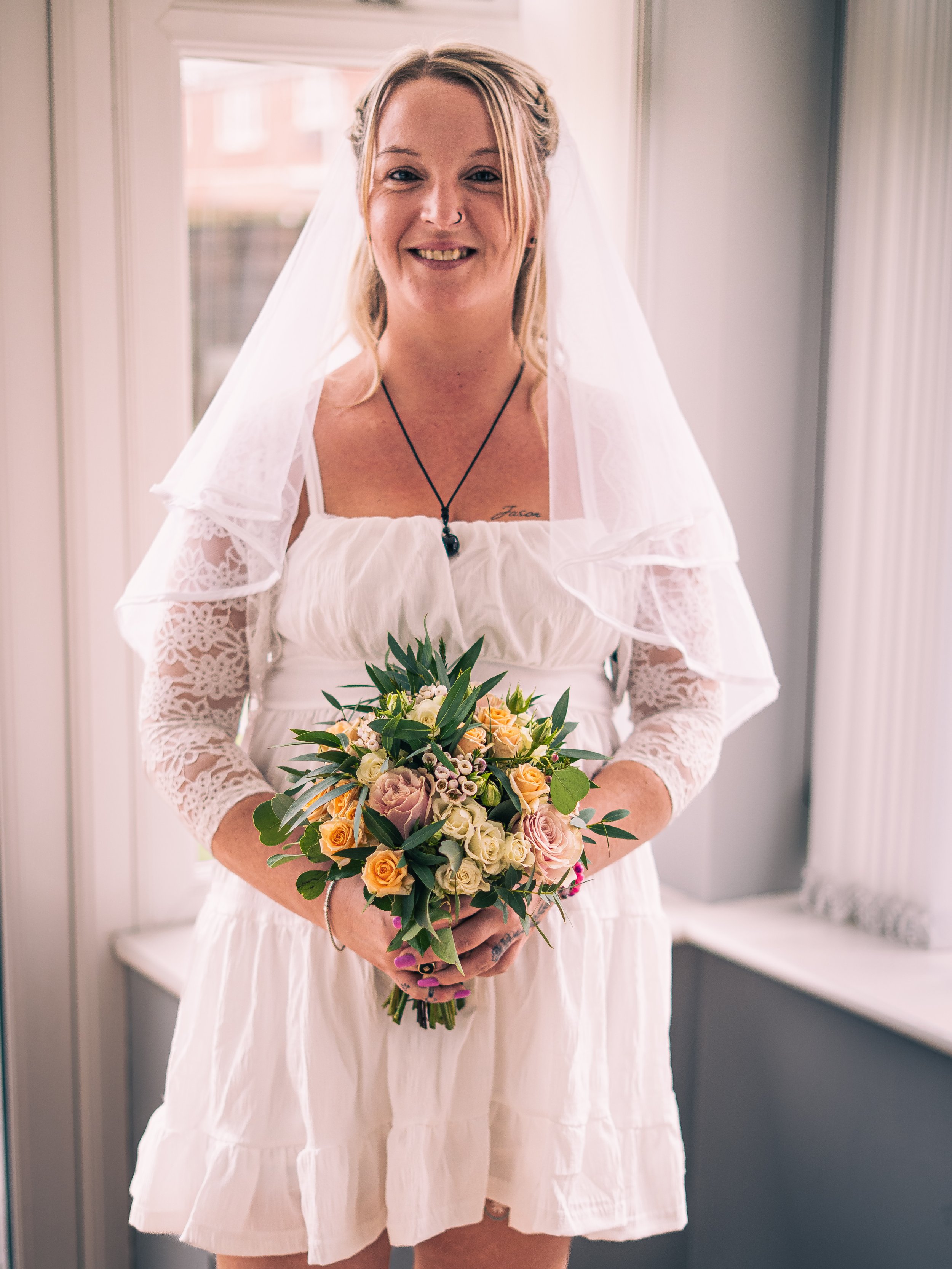 A smiling woman in a white dress holding a bouquet of flowers, standing in front of a window.