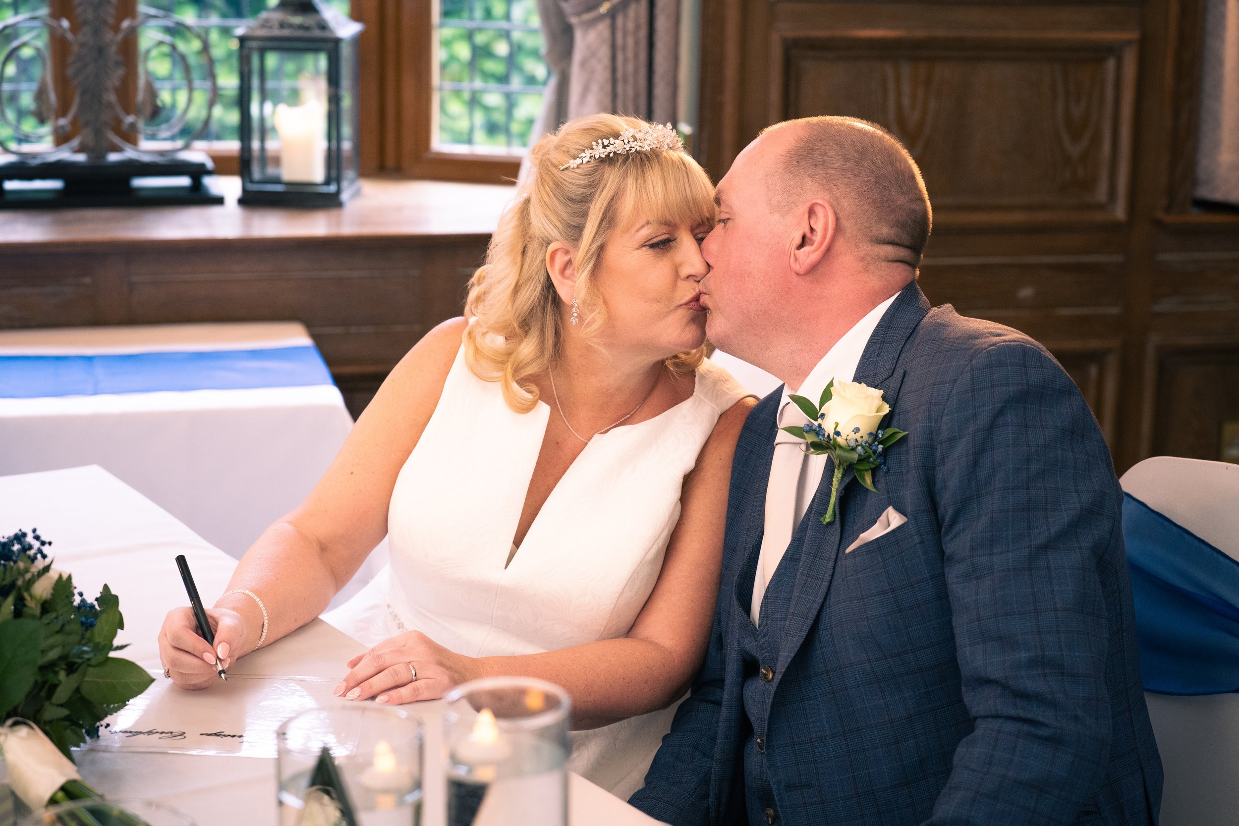 A bride and groom kissing at their wedding reception, with the bride writing on a guest book.