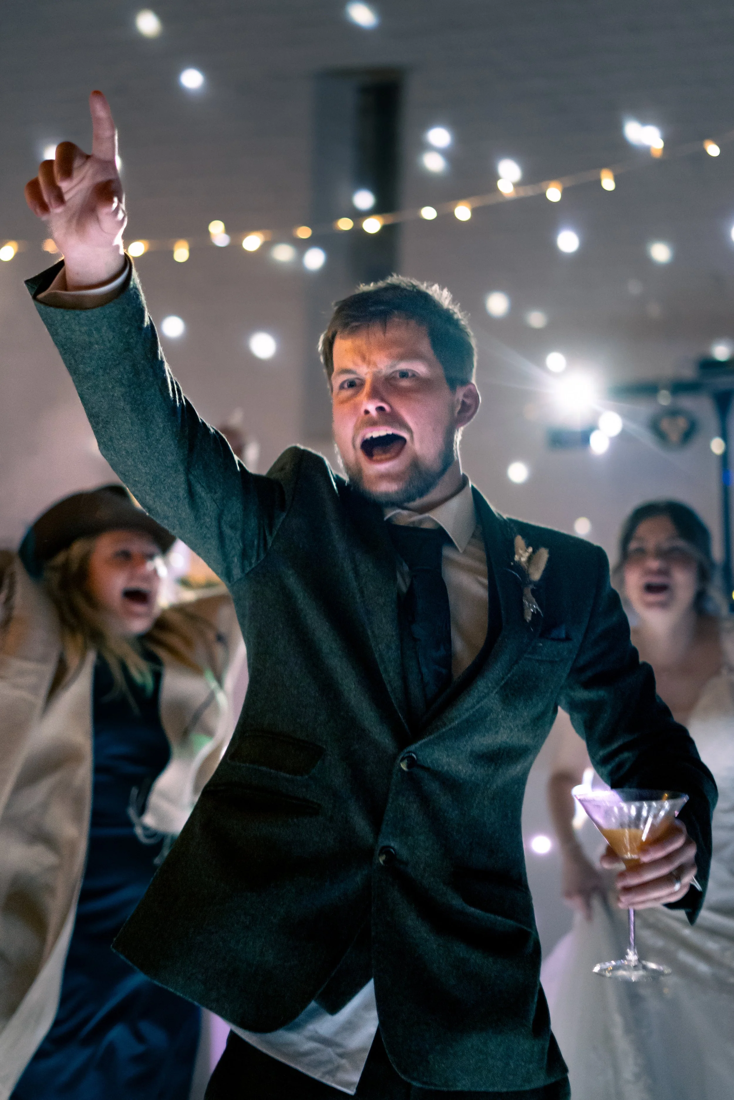 A man in a suit holding a martini glass, dancing or celebrating with others in the background at a party or wedding reception with string lights and a festive atmosphere.