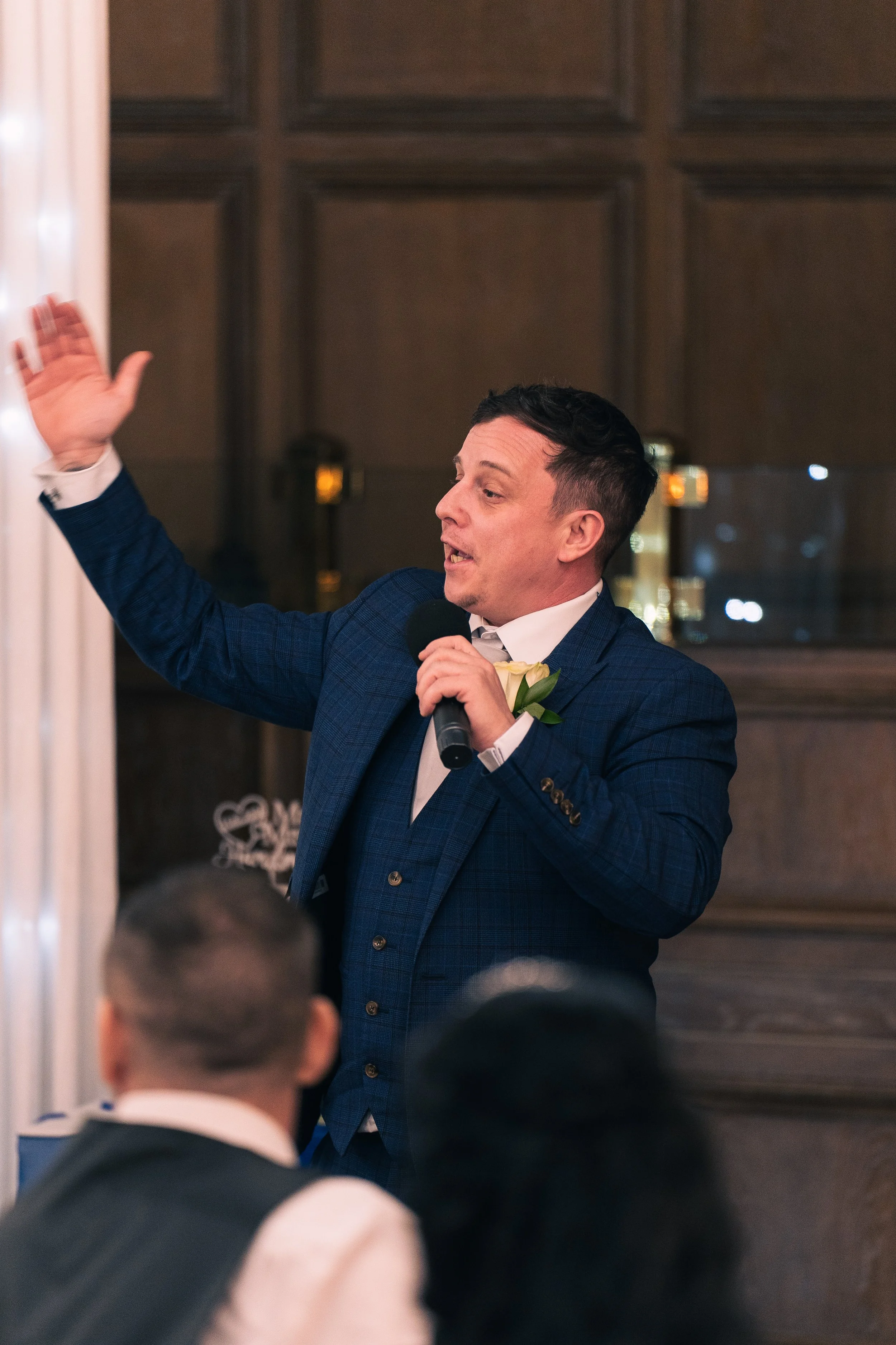 A man in a blue suit giving a speech at an event, holding a microphone in his right hand and raising his left hand.